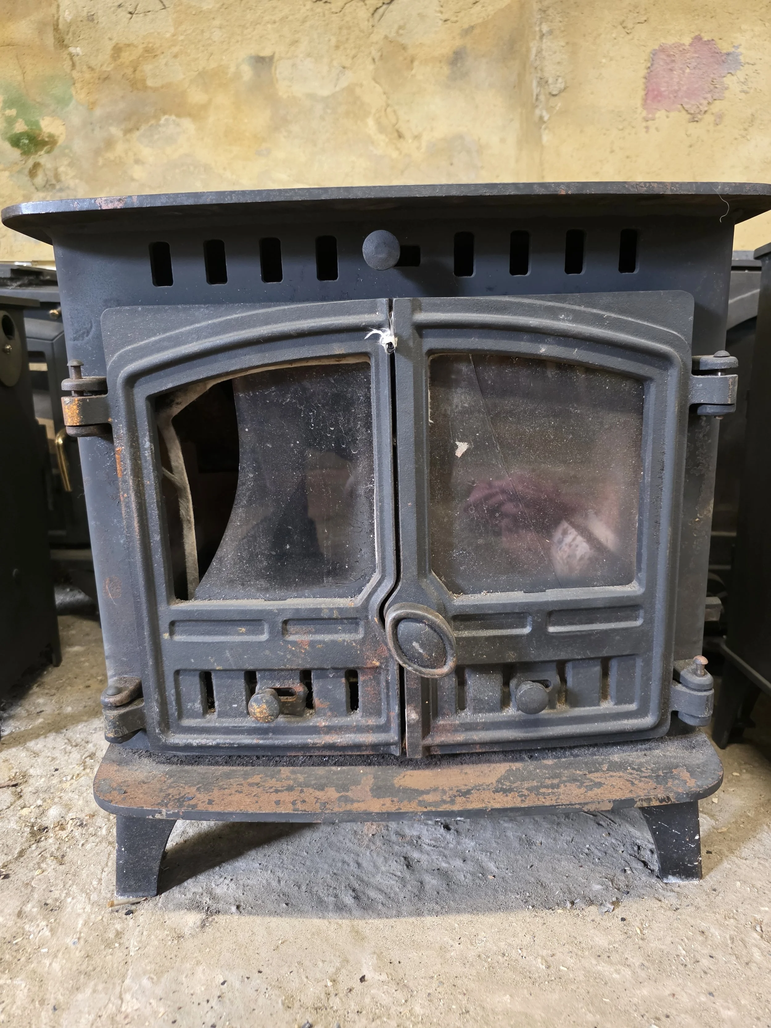 Front view of an old Herald Hunter 6kw before refurbishment  cast iron wood stove with double doors, sitting on a rusty metal base, in a rustic setting with a stained wall in the background.