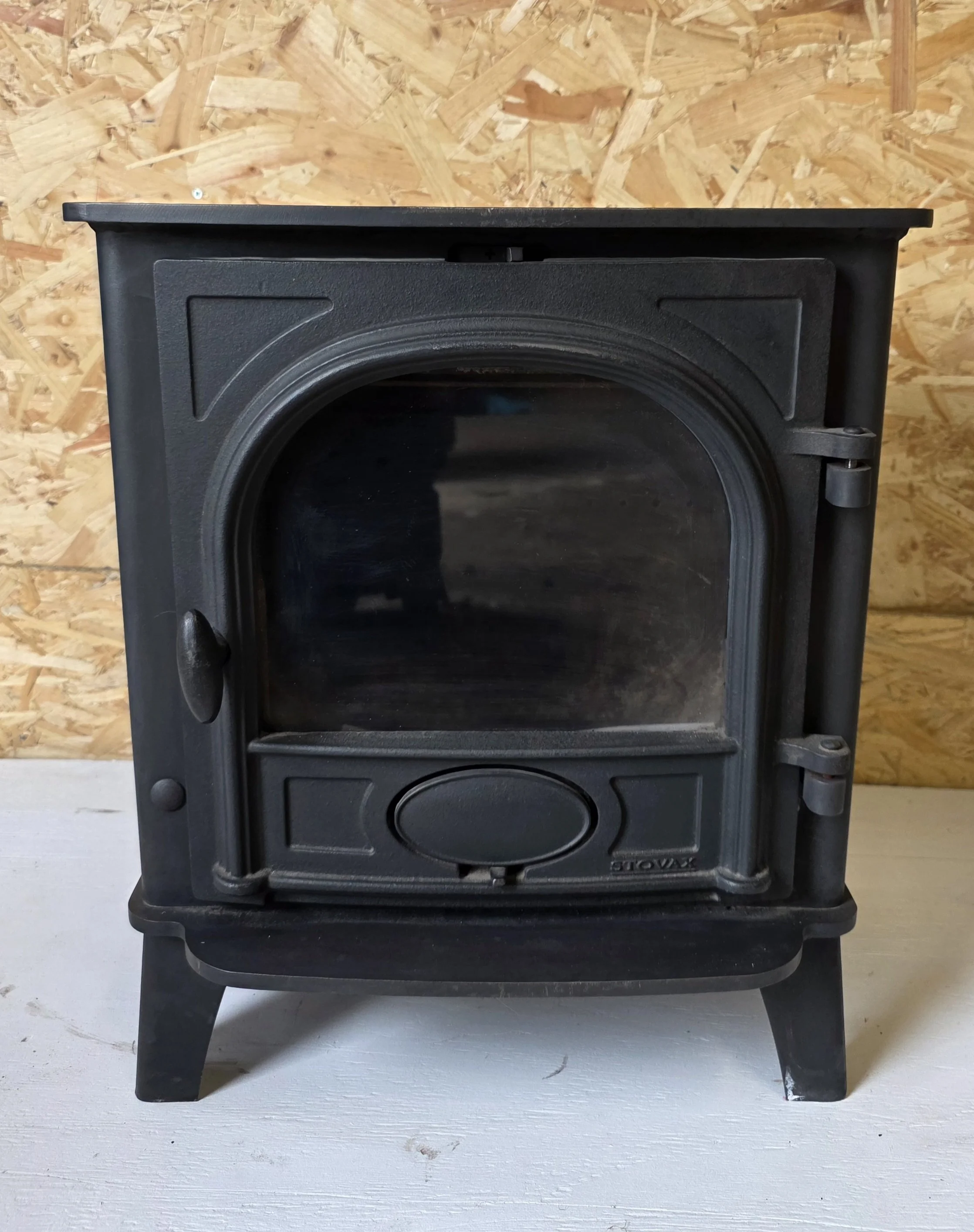 Black Stovax Stockton 5kw prior to refurbishment with a glass front door on a white wooden surface, with a wooden wall in the background.