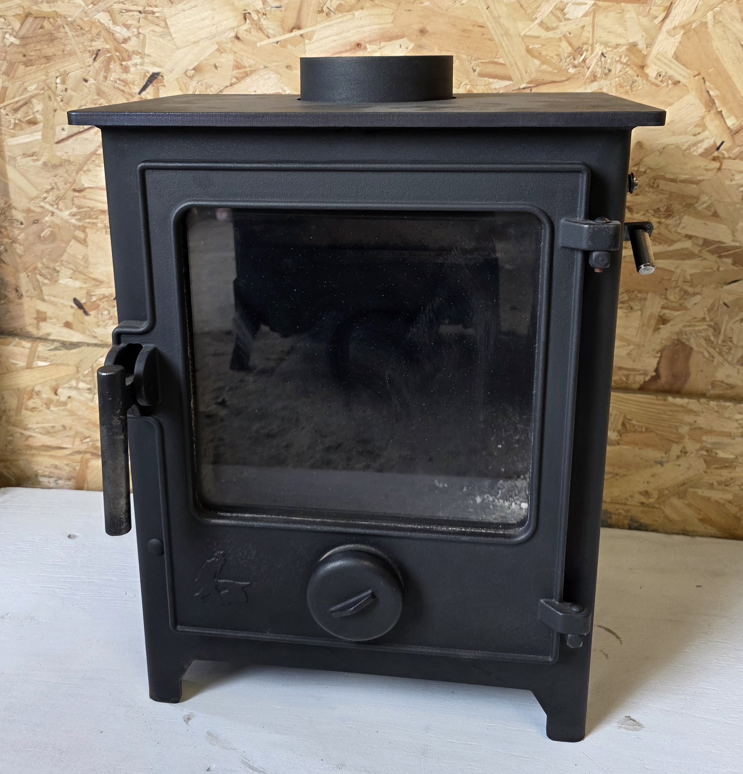A black Dean Forge Dartmoor 5 un refurbished wood-burning stove with a glass door, sitting on a white surface, against a background of unfinished wooden paneling.