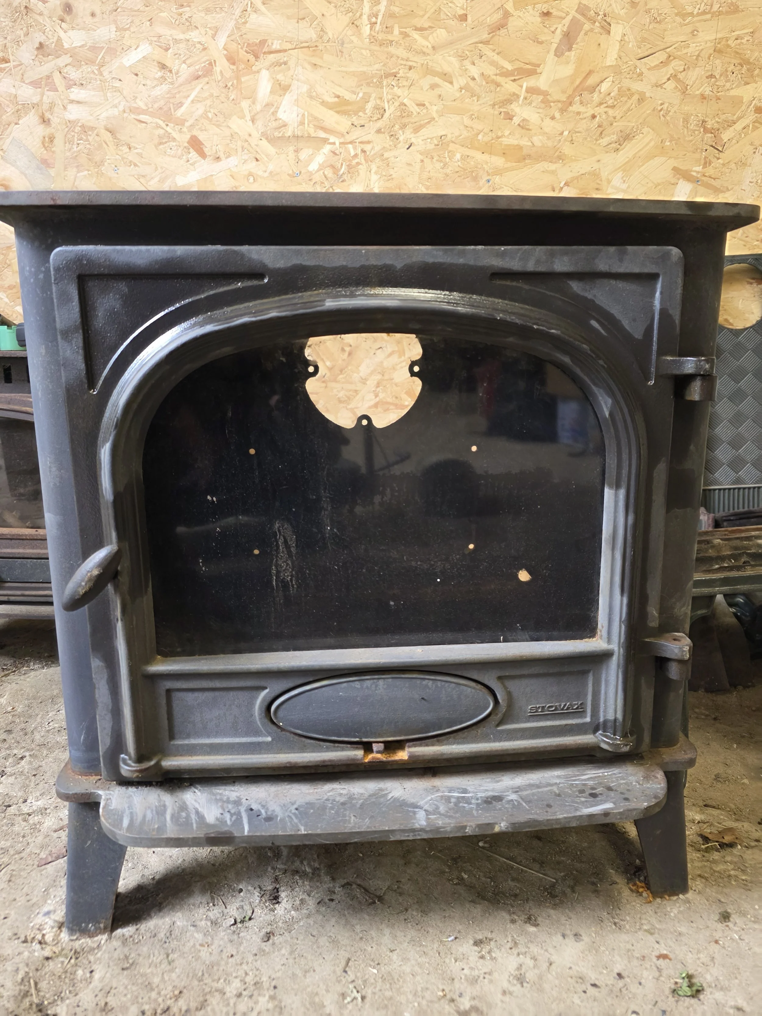 Front view of a black outdoor wood or pellet stove with a large glass door, sitting on a wooden surface in a workshop with a wood-paneled wall in the background.