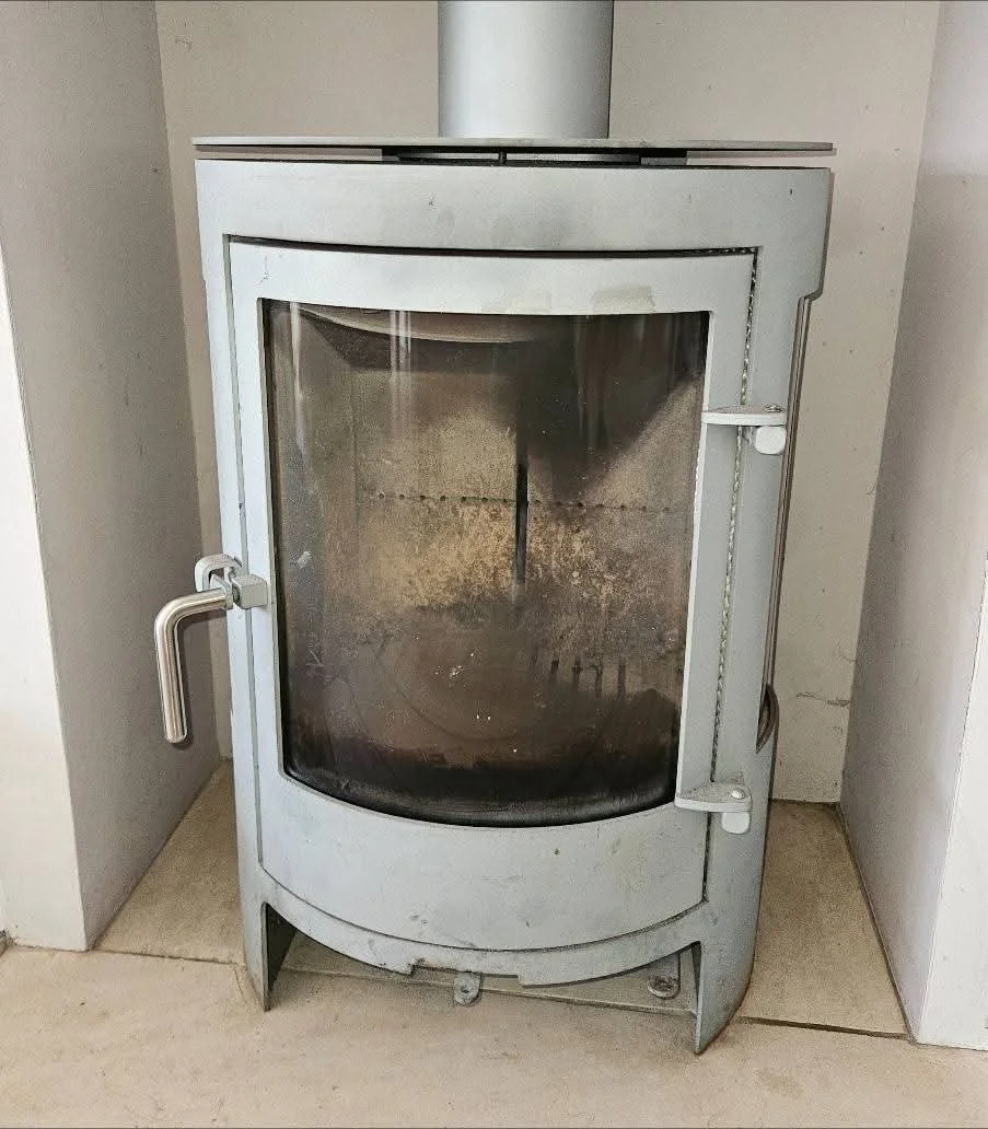 A vintage, metal wood stove with a curved glass door, dirty with soot inside, situated between two white walls.