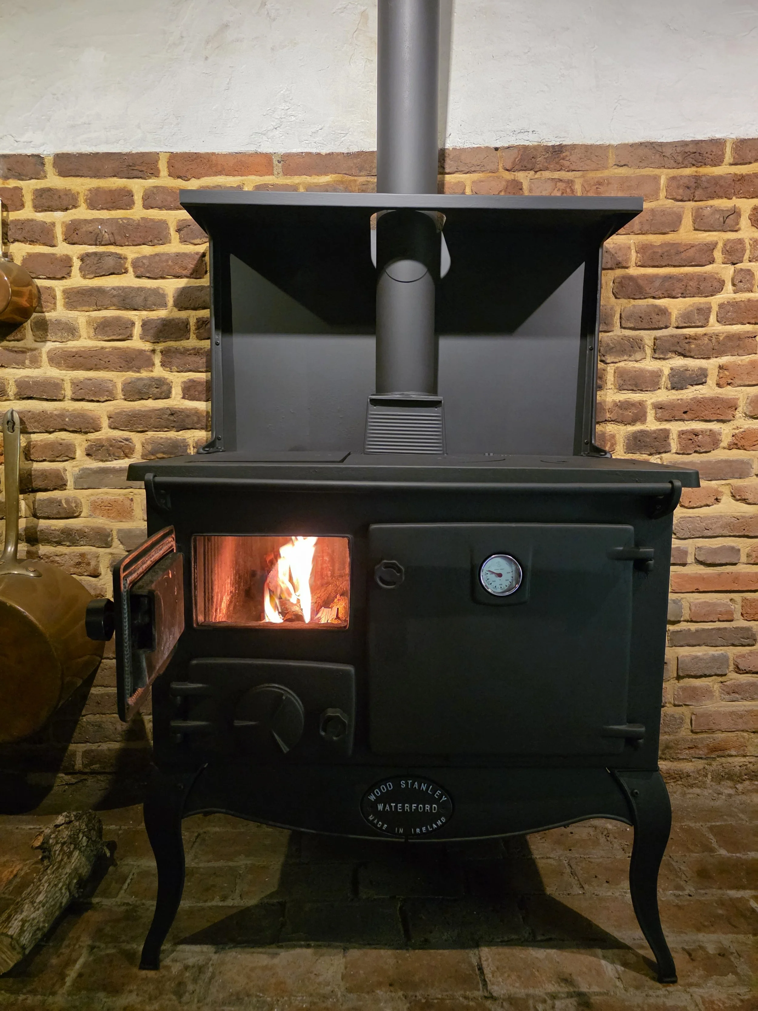 A black wood stove with a fire inside, set against a brick wall with a white upper section, and a chimney pipe extending from the stove to the ceiling.