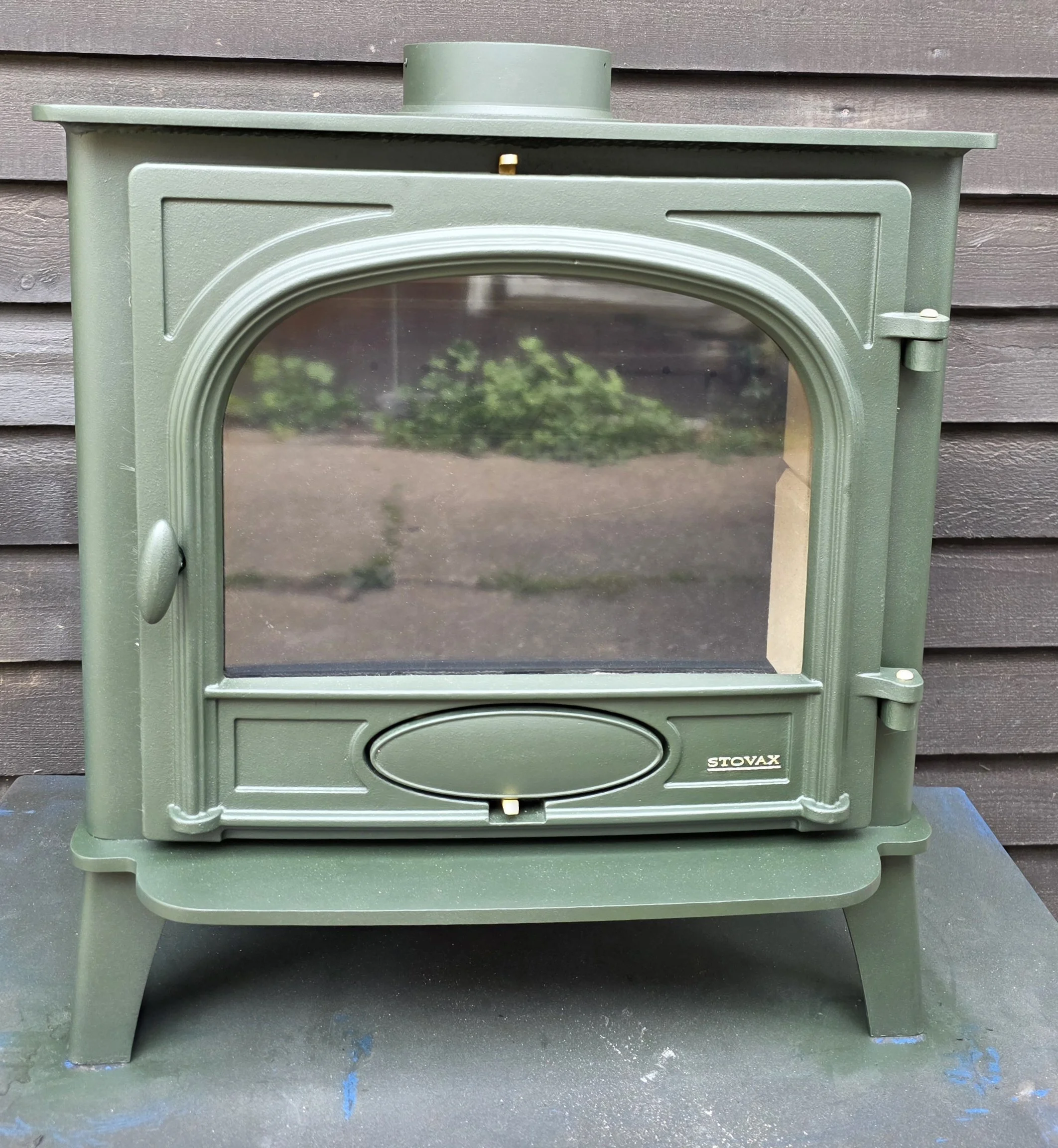 A green manual outdoor stove with a glass window on the door and metal handles, positioned in front of a wooden wall.