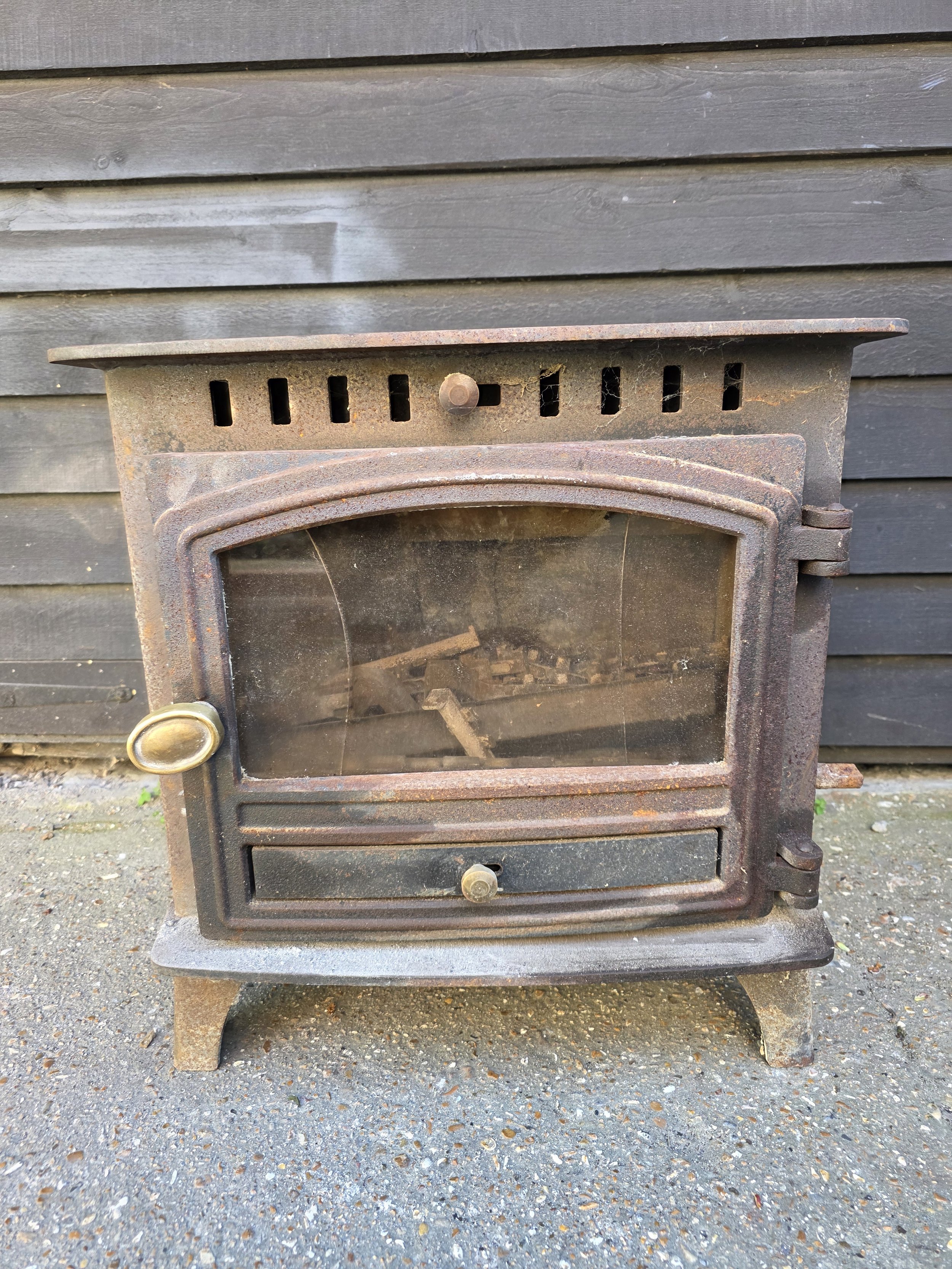 An old rusty outdoor wood stove with a glass door, set against a grey wooden wall, on a gravel ground.
