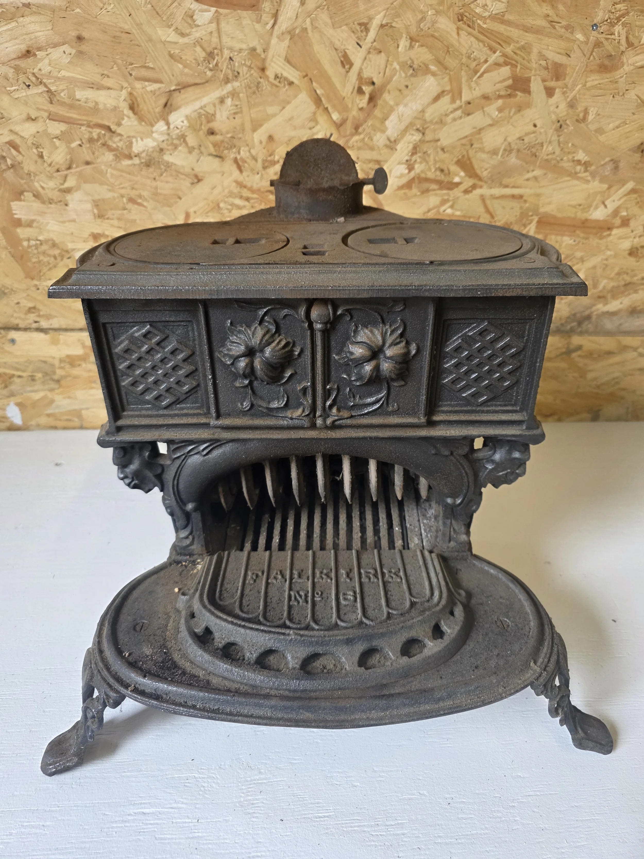 An antique cast iron stove from 1920's prior to refurbishment with decorative floral and geometric patterns, sitting on a white surface against a plywood wall.