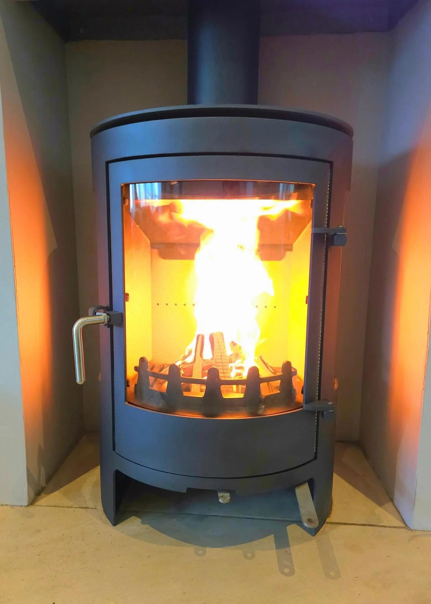 A modern black wood stove with glass door, burning bright orange flames inside, situated in a cozy corner on a beige tile floor.