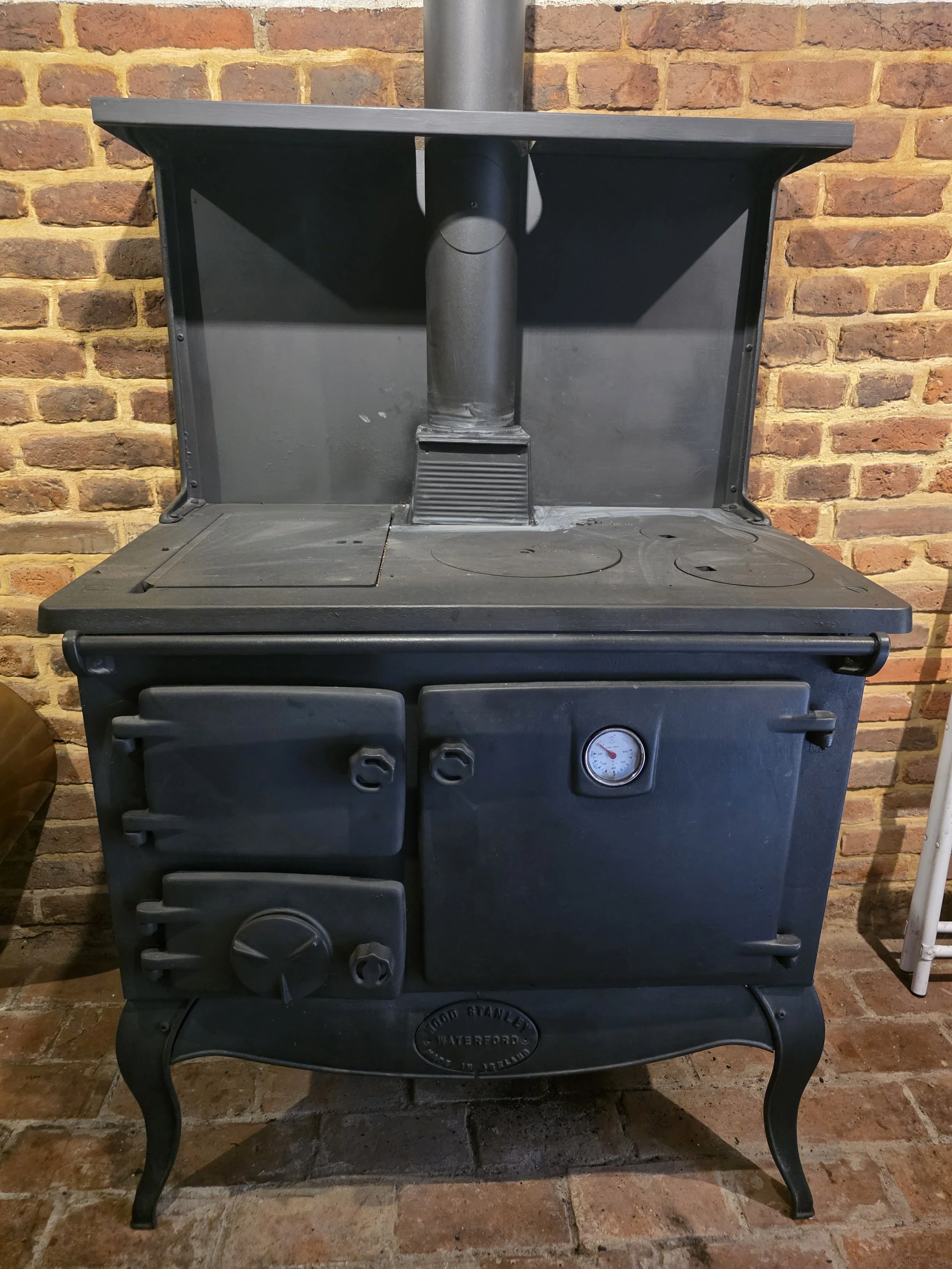 A vintage black cast iron stove with a round temperature gauge in a kitchen with a brick wall background.
