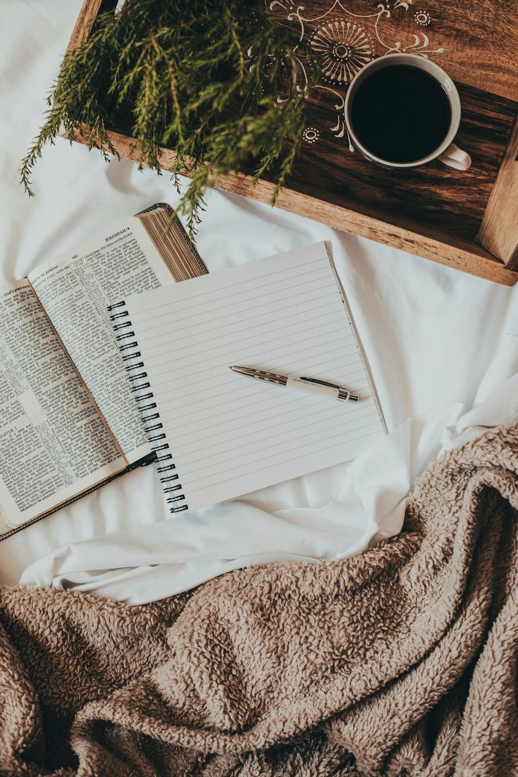 A cozy scene with a notebook and pen on a bed, a Bible open to Jeremiah 1, a cup of coffee on a wooden tray, and a green plant, all surrounded by a soft, beige blanket.