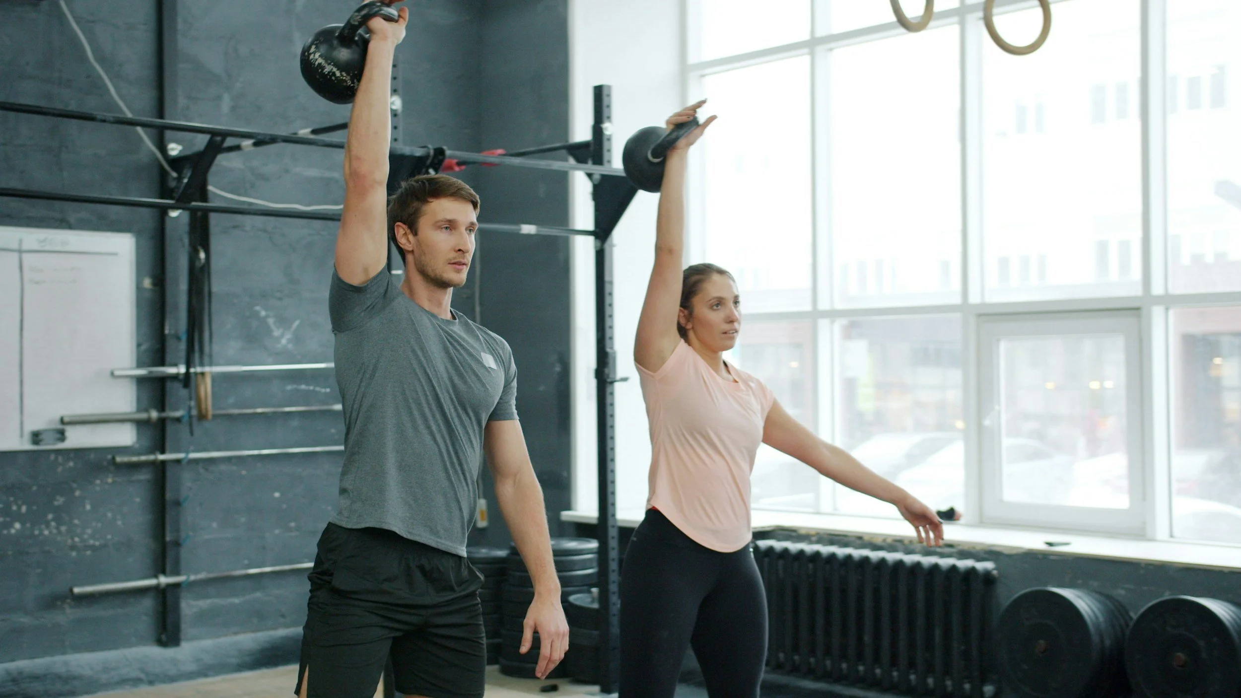 A man and woman exercising with kettlebells in a gym, raising them above their heads in a workout class.
