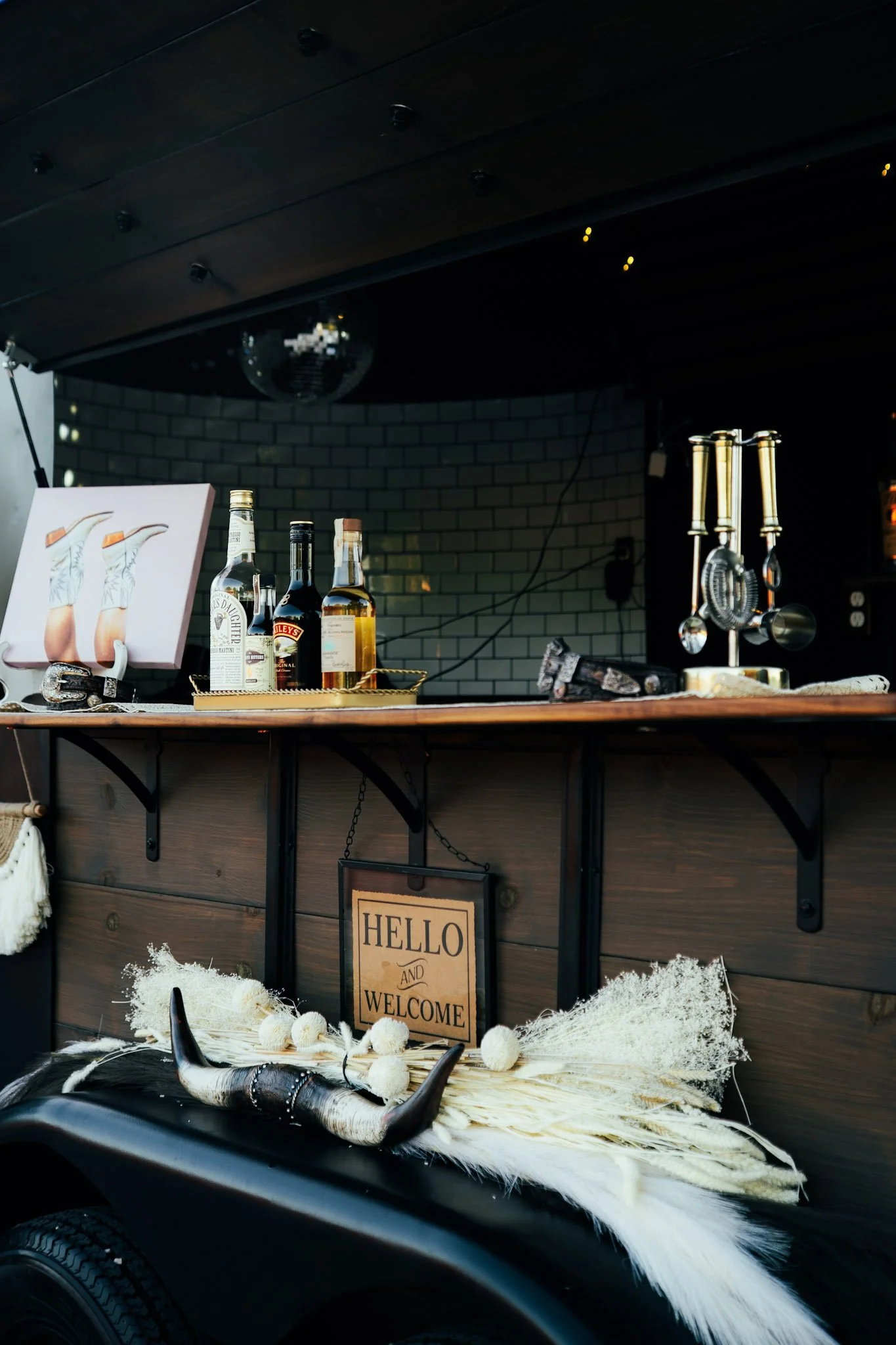 Bar cart with liquor bottles, a sign reading 'Hello and Welcome,' decorative horns, and dried plants on the lower shelf, with a white brick wall in the background.