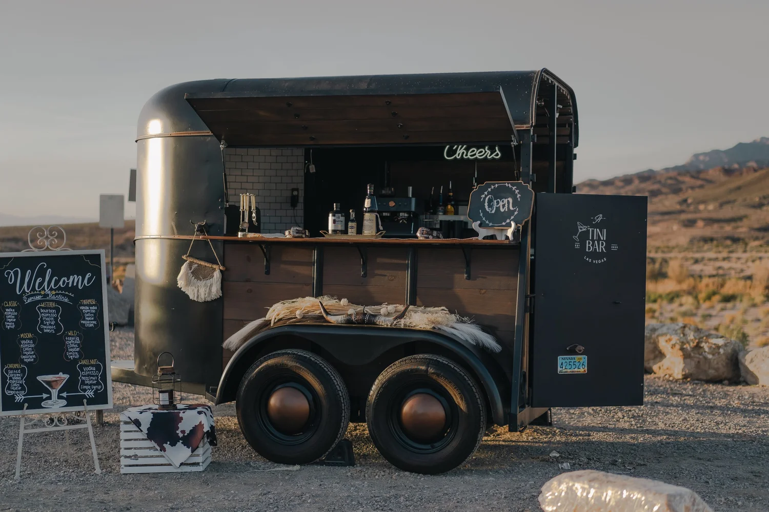 A Tini Bar's vintage horse trailer bar with a welcome sign and Cheers neon, set up in the Nevada desert at dusk