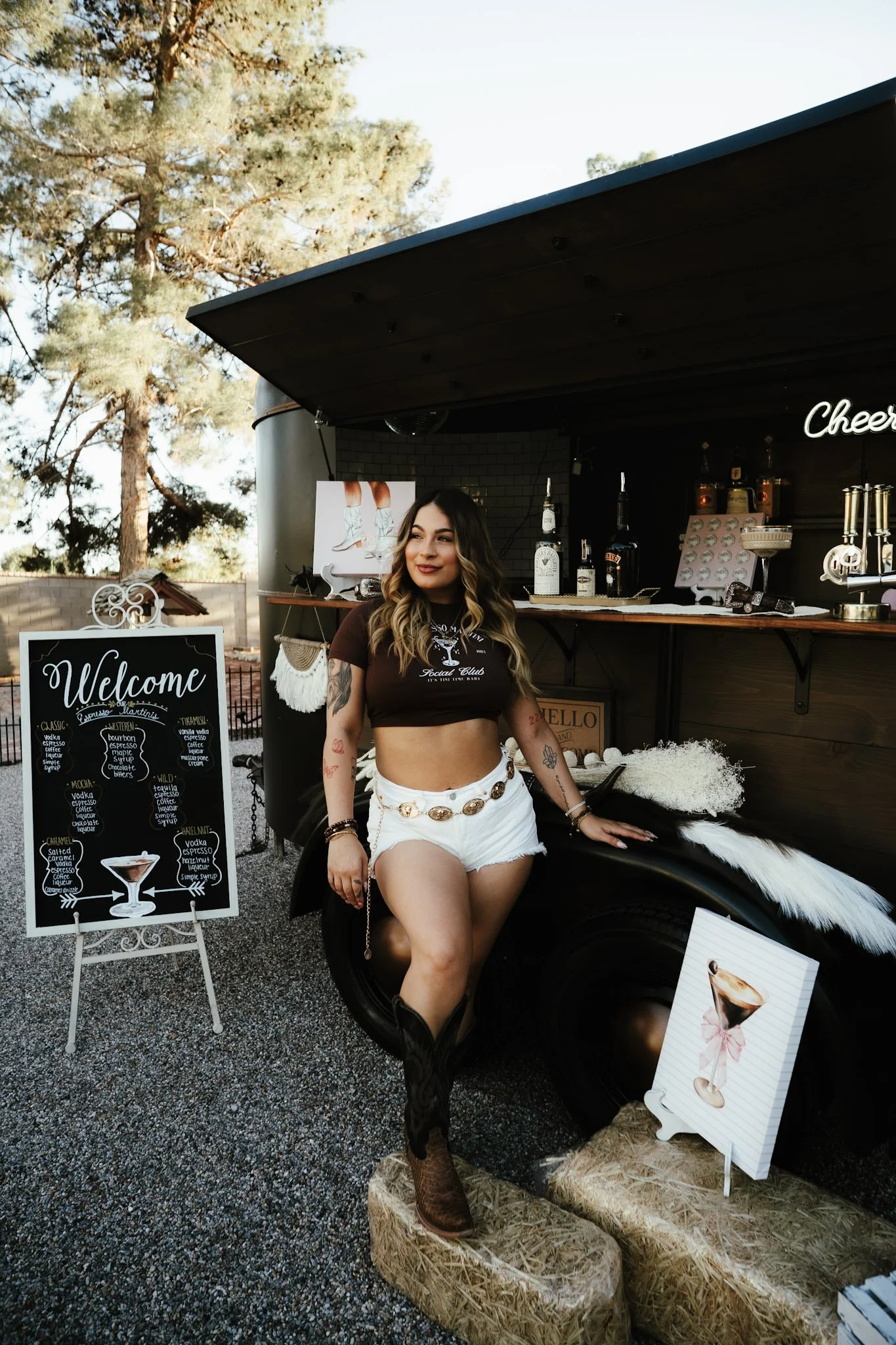 Woman in a black crop top and white shorts standing next to a champagne bottle sign at an outdoor bar with trees in the background.