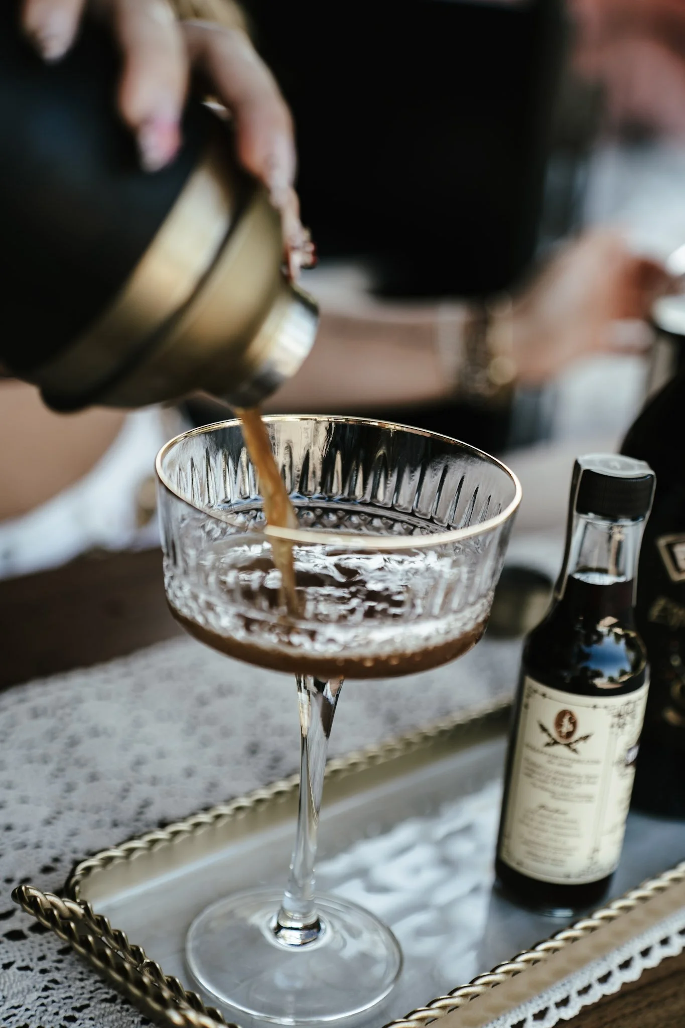 A person pouring a dark liquid, likely coffee or a cocktail, into a glass with a textured pattern on a silver tray that also holds a small bottle of dark syrup or liqueur.