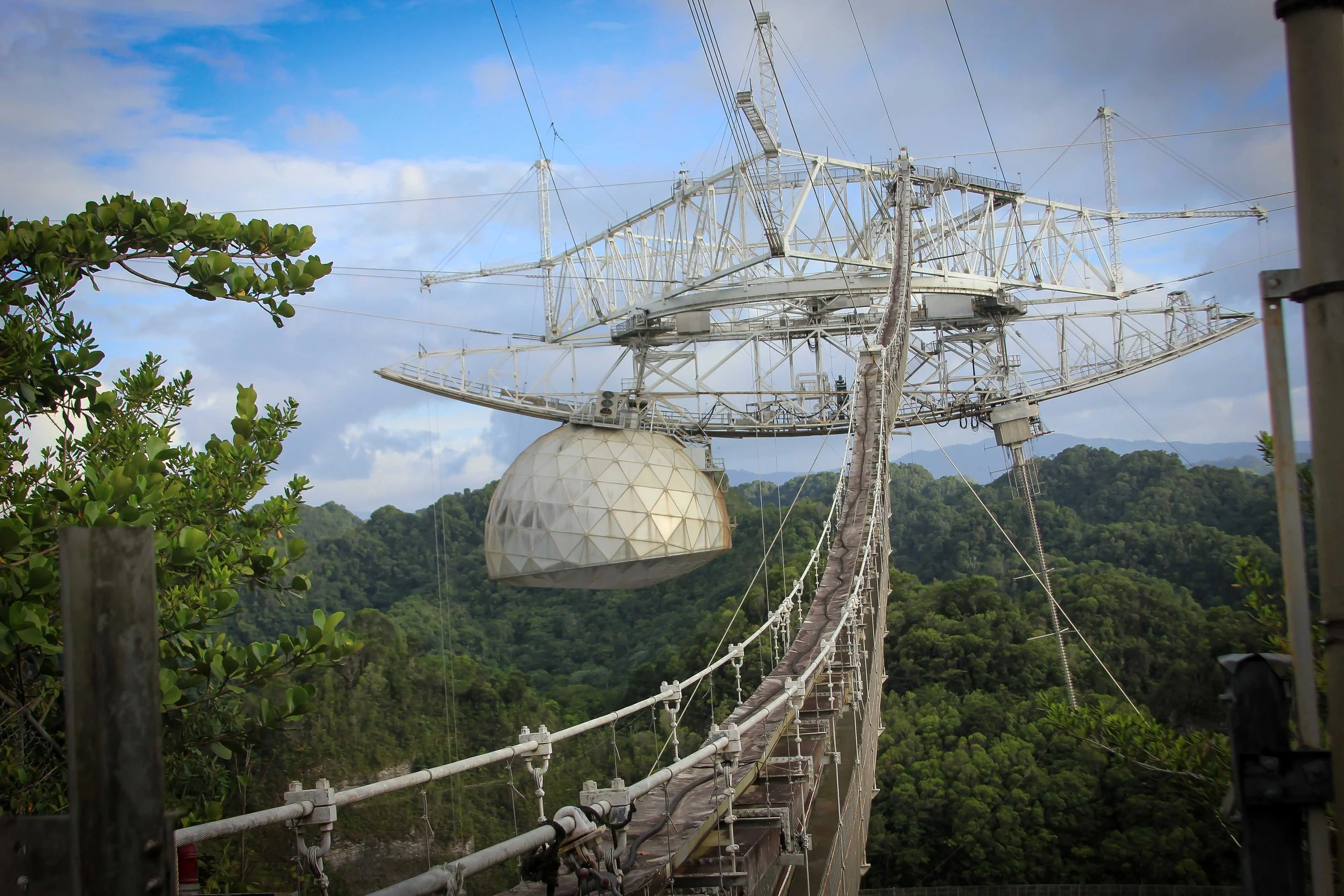 A large radio telescope structure with a spherical receiver in a forested mountainous area under a partly cloudy sky.
