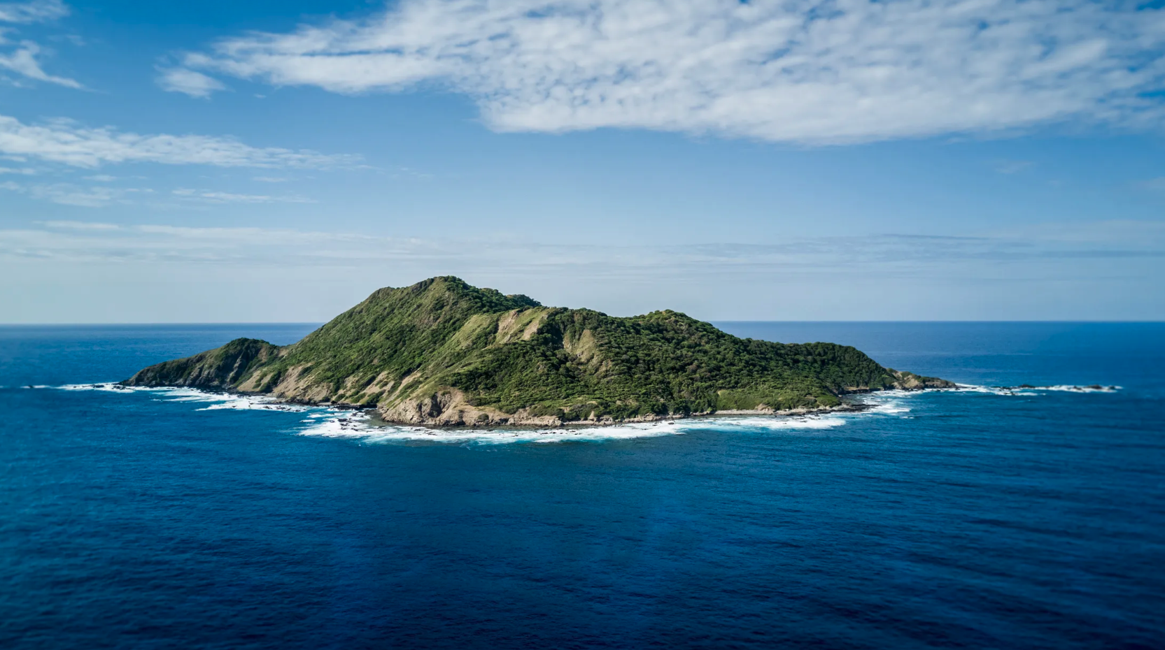 A lush green island with hilly terrain surrounded by deep blue ocean waters under a partly cloudy sky.