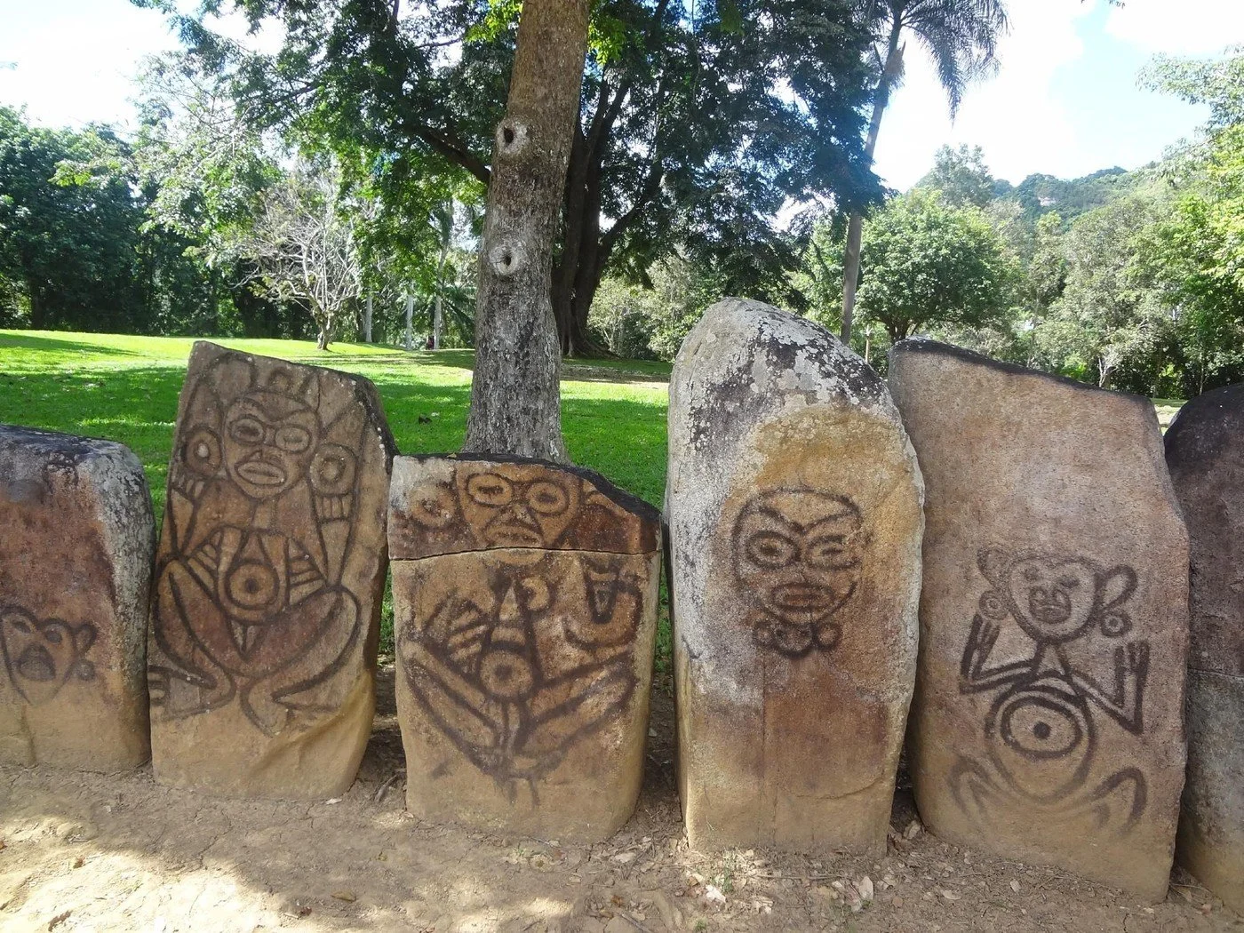Four large stones with ancient tribal carvings depicting human figures, in a grassy outdoor area with trees and a clear sky.