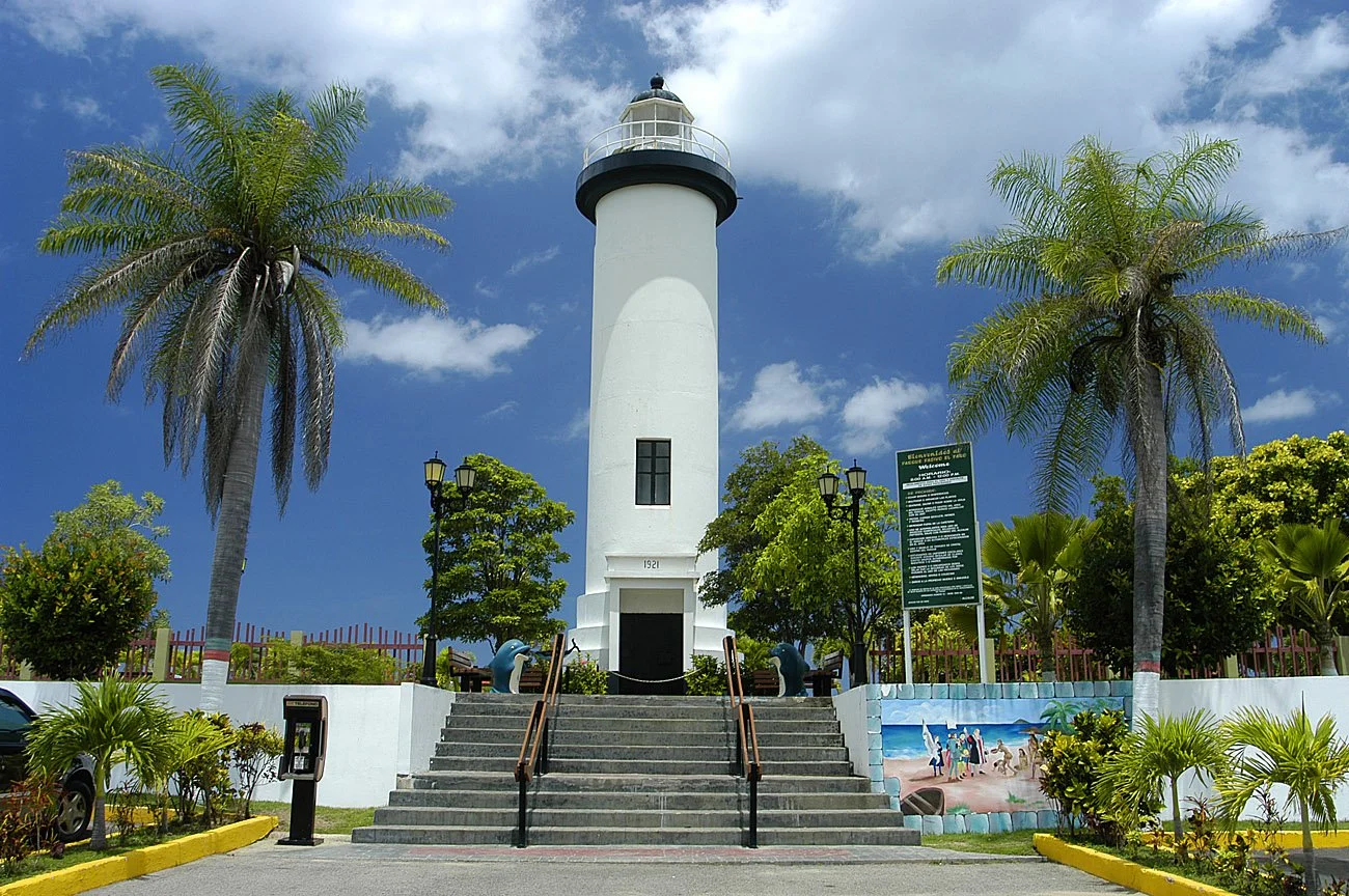 White lighthouse with a black top on steps surrounded by palm trees and greenery, under a partly cloudy sky.