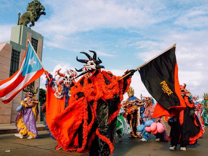 People dressed in colorful traditional costumes and masks participating in a parade, holding flags, with a monument and a lion sculpture in the background.
