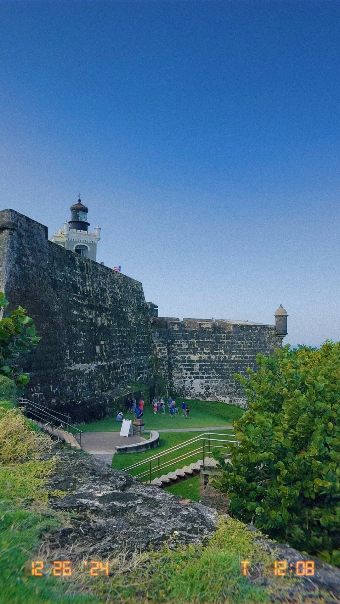 Historic fort with stone walls and a lighthouse on top, green landscaped area with a group of people walking, blue sky.