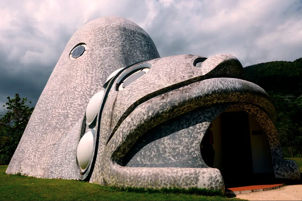 A large building designed to look like a stylized face with rounded features, large circular windows for eyes, and a curved entrance resembling a mouth, set outdoors with cloudy skies and greenery in the background.