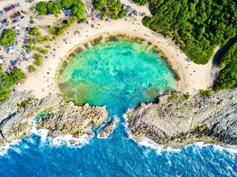 Aerial view of a small bay with turquoise water, sandy beach, rocky cliffs, surrounded by green trees, and a parking lot with cars nearby.