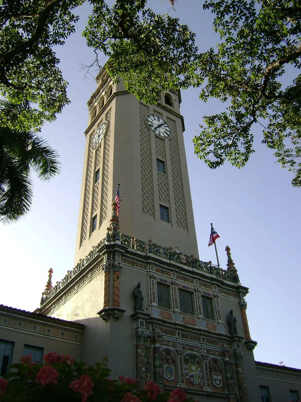 A tall clock tower with a decorative base, flags, and surrounded by green trees against a clear blue sky.