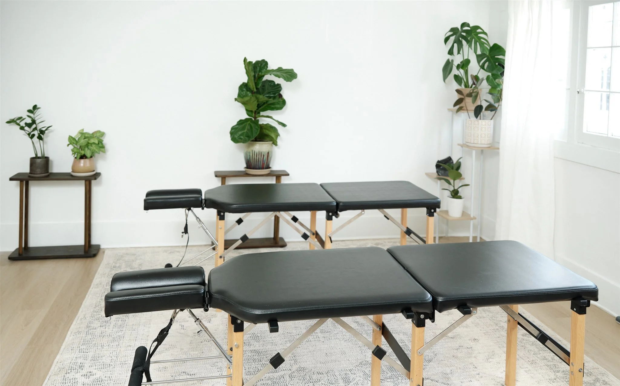 A room with chiropractic tables, several potted plants on small tables and shelves, and white walls and curtains.