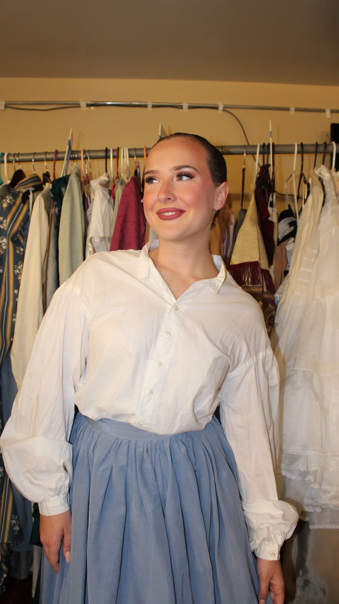 Person wearing a white blouse and blue skirt in a dressing room with hanging clothes.