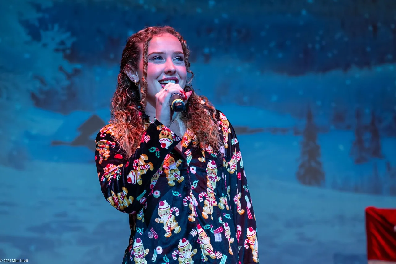 A person with curly hair singing into a microphone wearing a Christmas-themed shirt featuring gingerbread men and candy canes, standing in front of a snowy winter background.