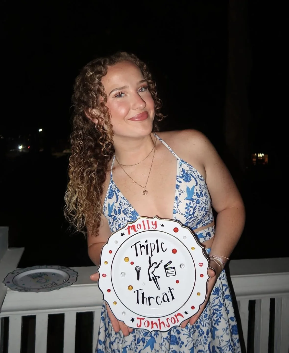 A person with curly hair in a blue and white dress holds a decorative plate labeled 'Triple Threat Molly Johnson' with theater-related symbols.