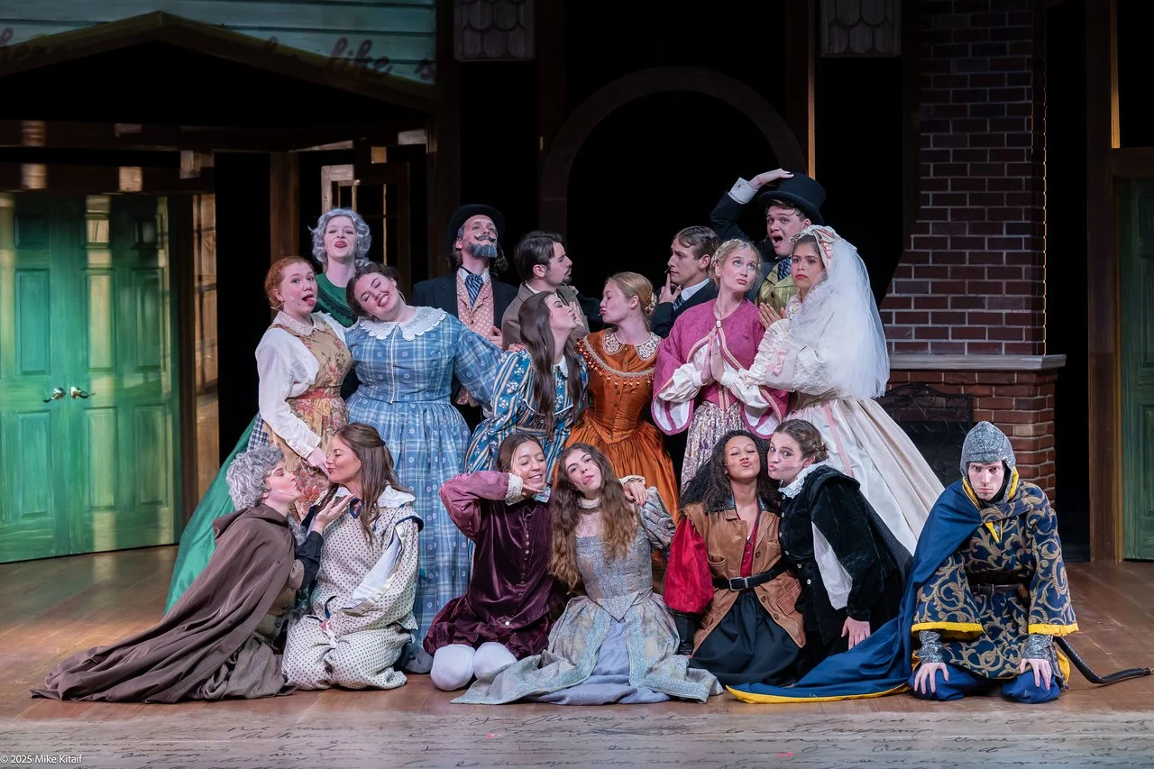 A group of people in period costumes poses dramatically on stage, suggesting a theatrical performance or play. The backdrop includes elements of a building with green doors and a brick wall. The participants wear various types of historical clothing, including dresses and medieval armor.