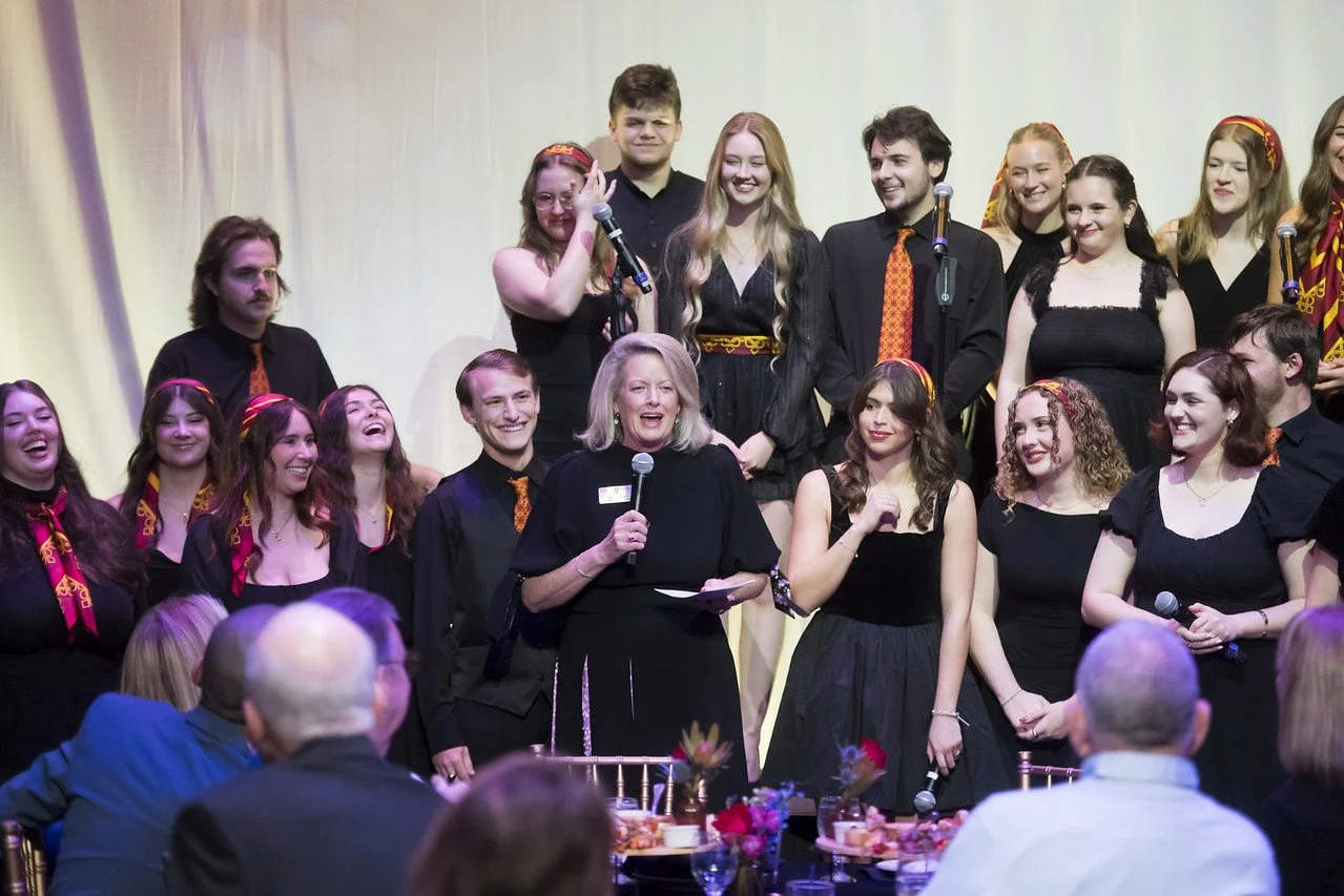 A group of choir singers dressed in black with red and gold accents, standing on a stage. A woman holding a microphone speaks to an audience. The setting appears to be a formal event with decorated tables and attendees seated.