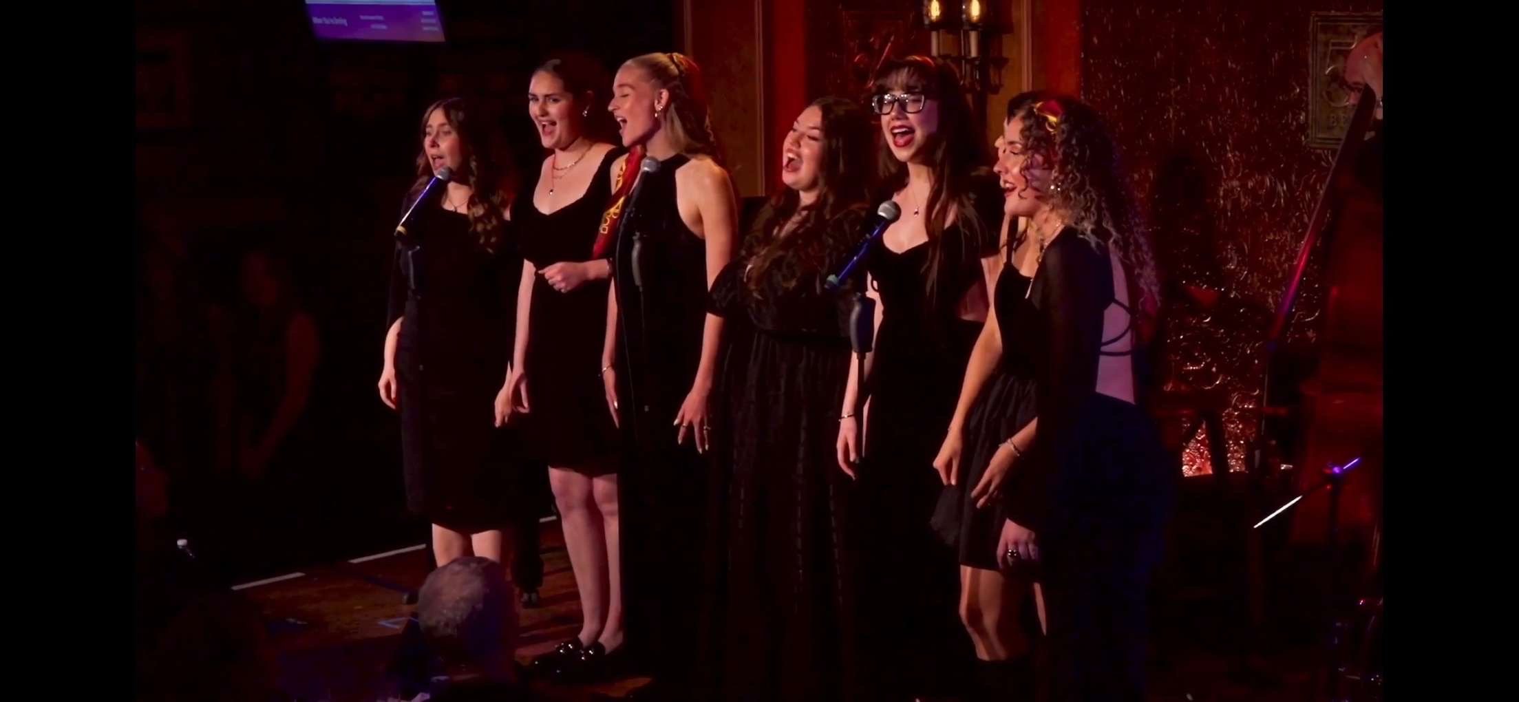 Group of six women singing on stage in black dresses