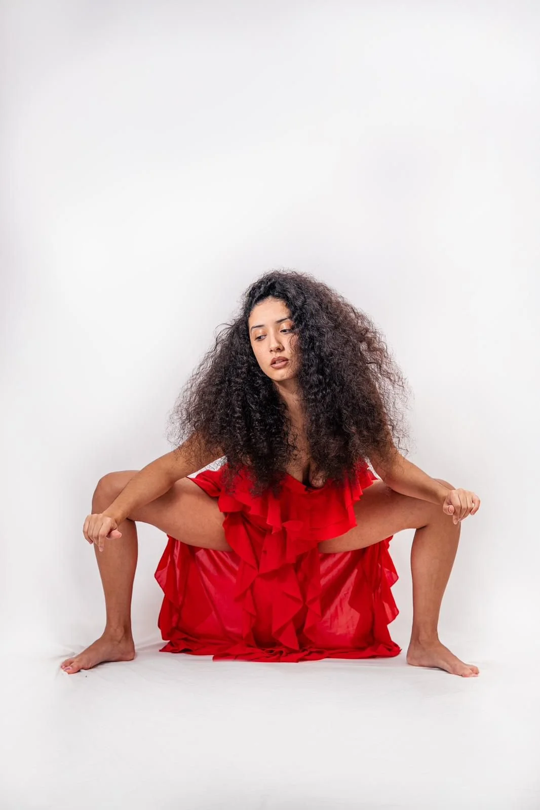 A woman with curly hair in a red dress squat with her arms bent, set against a plain white background.