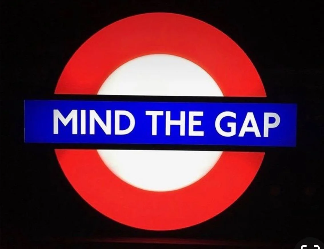 A circular sign resembling a London Underground logo with a red outer ring, a white inner circle, and a blue rectangle across the center displaying the text "MIND THE GAP" in white letters.