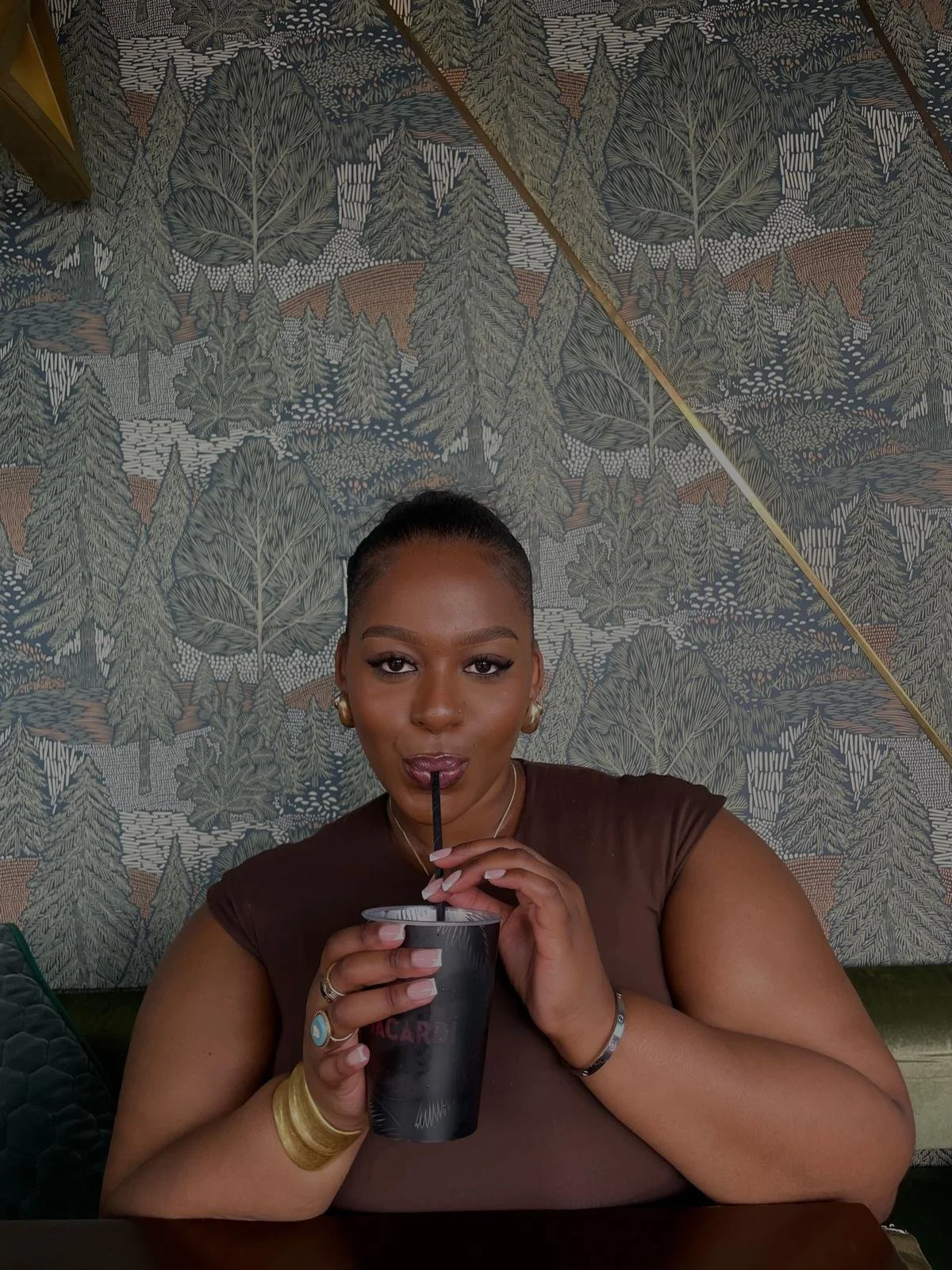 A woman with dark hair in a bun, wearing gold earrings and a brown top, is sipping a drink through a straw from a black cup, sitting in front of a wall with a forest pattern wallpaper.