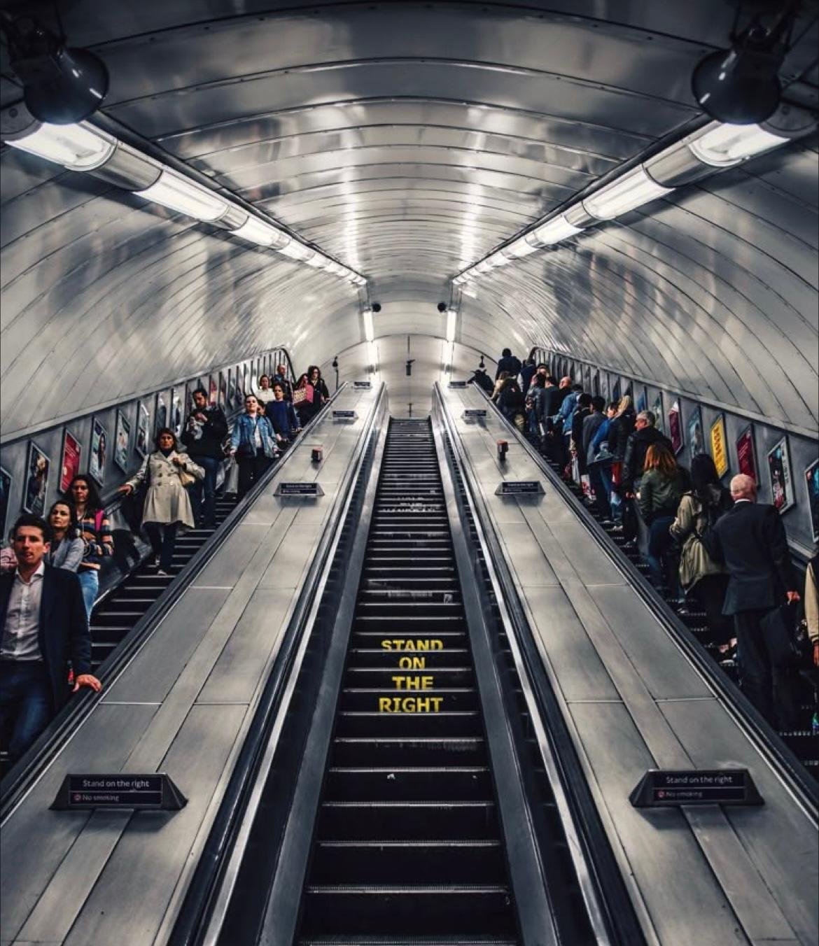 An underground metro station with two escalators going up, crowded with commuters on either side. The tunnel has a metallic, curved ceiling with bright fluorescent lights. Yellow text on the escalator reads, 'STAND ON THE RIGHT.'