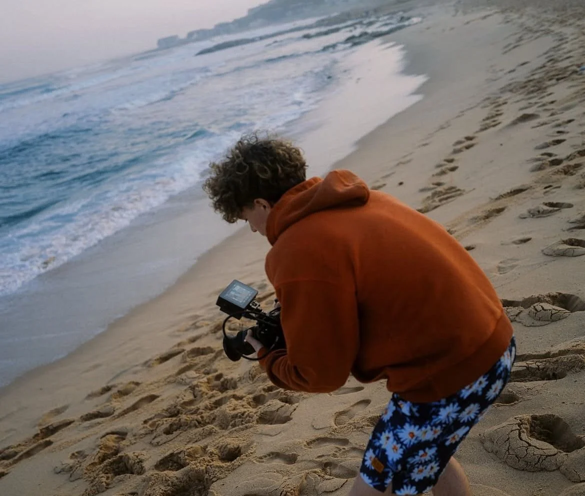 A person with curly hair wearing an orange hoodie and floral shorts is on a sandy beach, holding a camera while looking at the ocean and waves during sunset or sunrise.