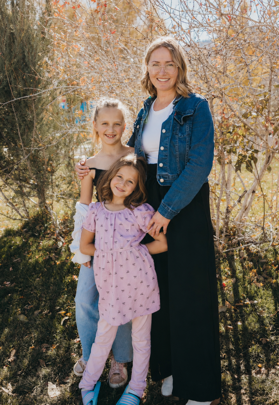 A woman with glasses and short wavy hair standing outdoors with two young girls, all smiling, surrounded by autumnal trees with orange and red leaves.