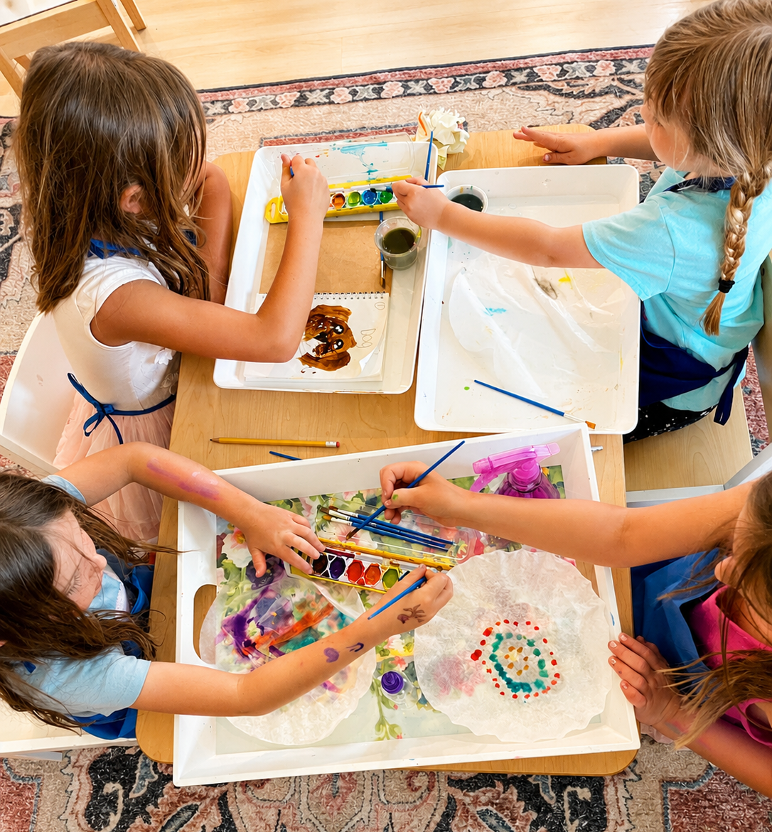 Four children sitting at a table painting with watercolors and brushes, creating art on paper.