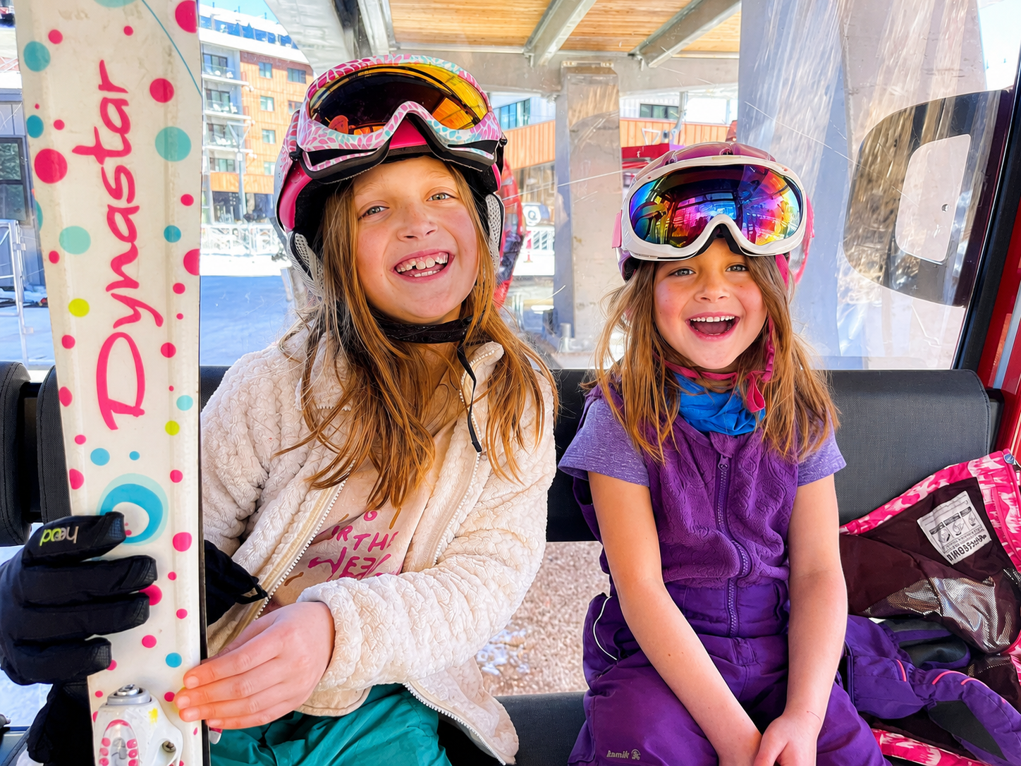 Two young girls in ski gear smiling while sitting inside a ski lift cabin, with snow, buildings, and a blue sky visible outside.