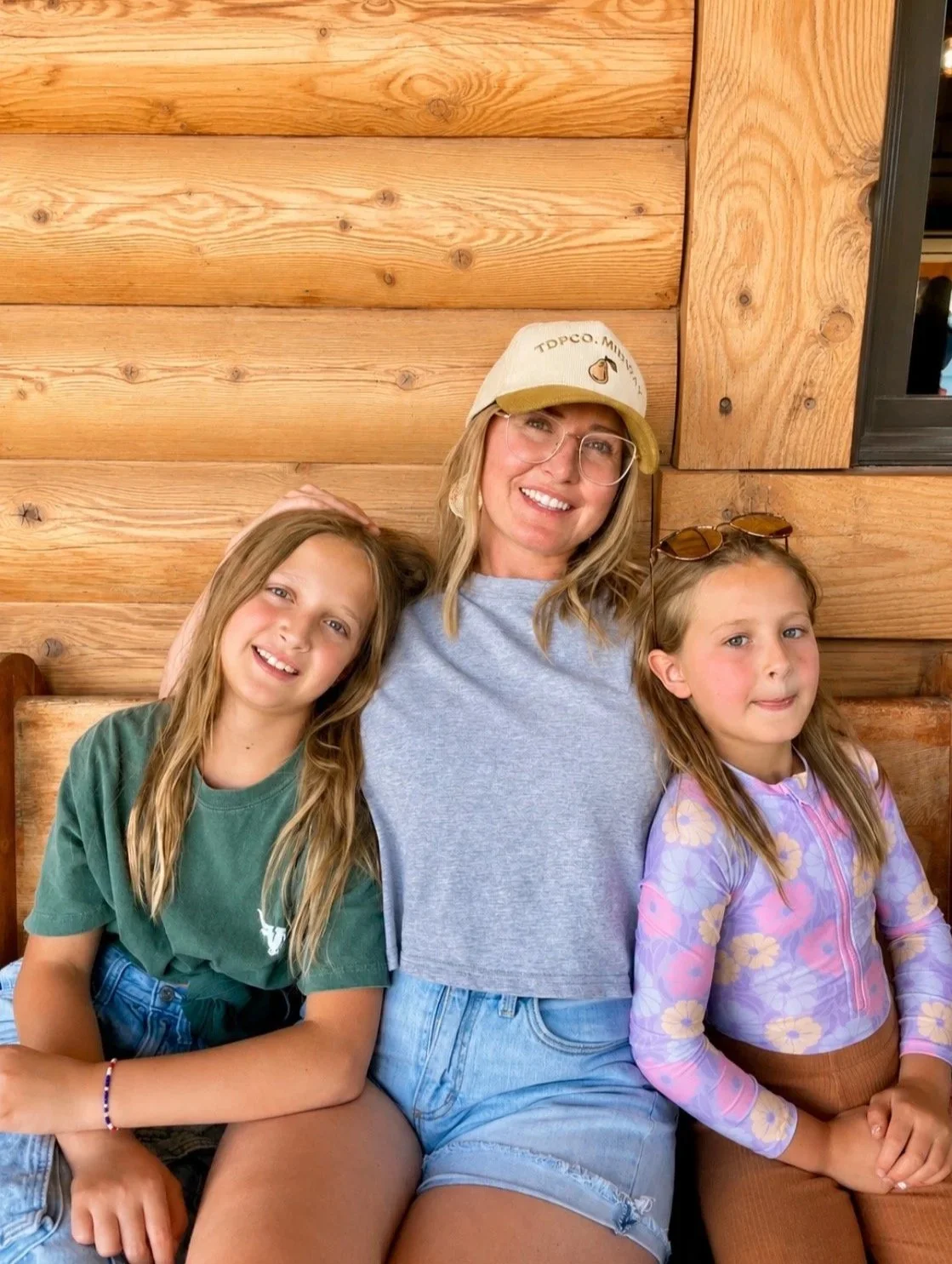 Three people, a woman and two girls, sitting together in front of a wooden wall. The woman is wearing a baseball cap, glasses, and a gray T-shirt. The girl on the left has long blonde hair, a green T-shirt, and a colorful bracelet. The girl on the right has long blonde hair, a purple floral jacket, and sunglasses on her head. They are smiling and appear to be enjoying each other's company.