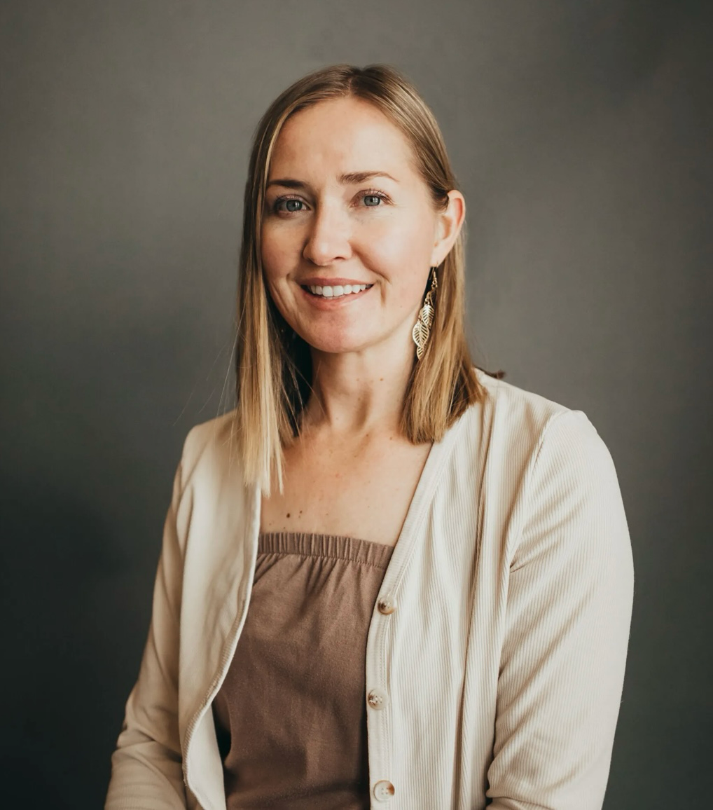A woman with straight blonde hair wearing earrings, a brown top, and a cream-colored cardigan smiling against a dark gray background.