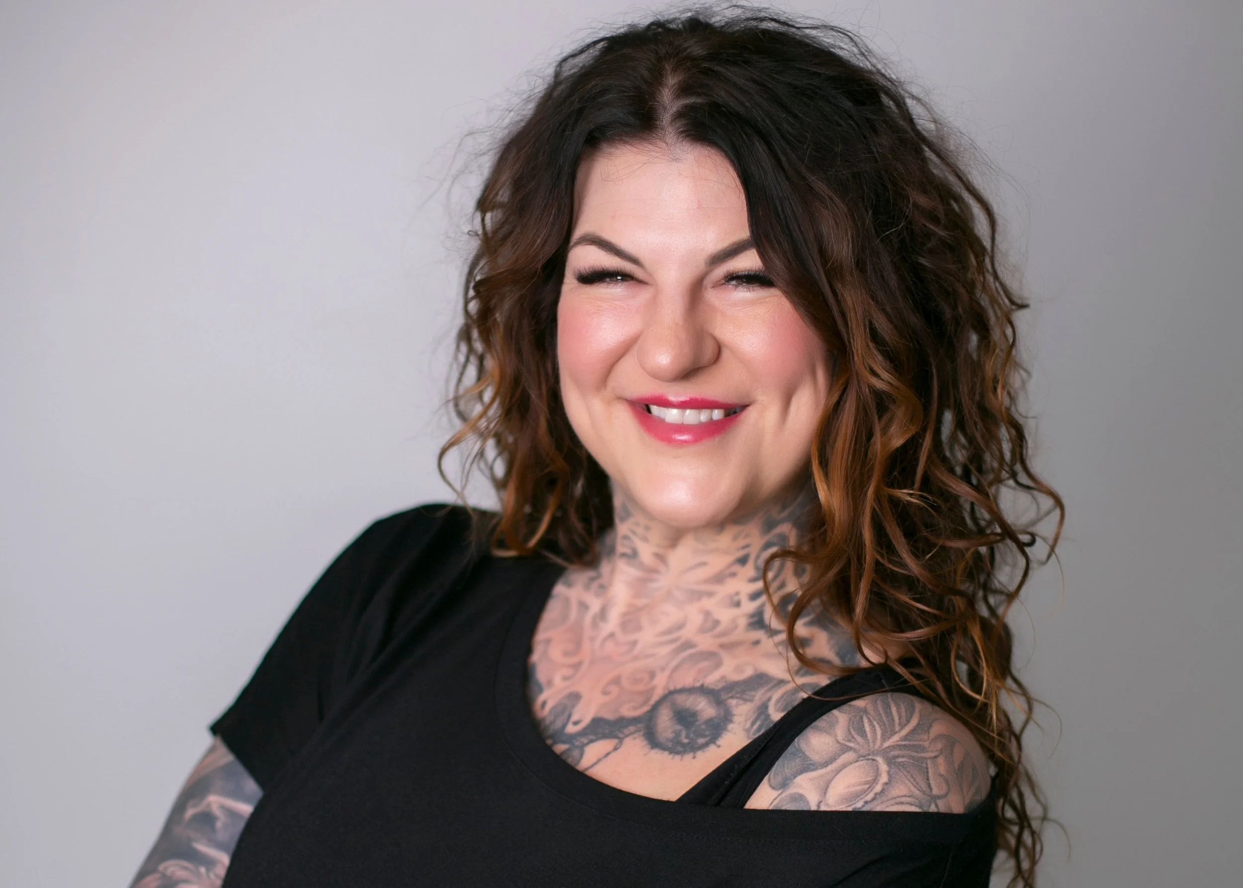Smiling tattooed woman with long wavy brown hair wearing a black top against a gray background