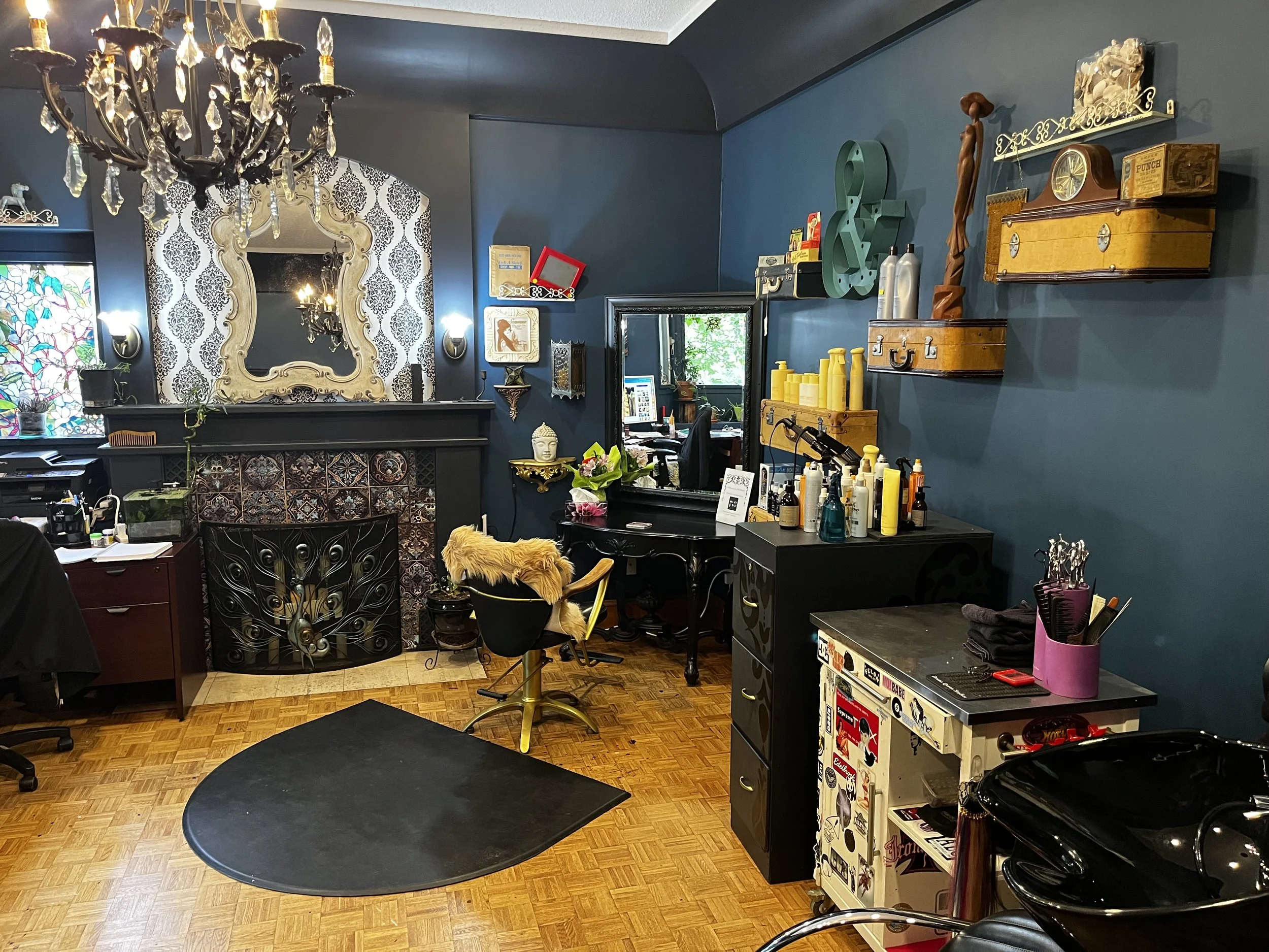 Vintage-inspired hair salon interior with ornate chandelier, decorative fireplace, styling chair, mirrors, hair products, and antique wall decor