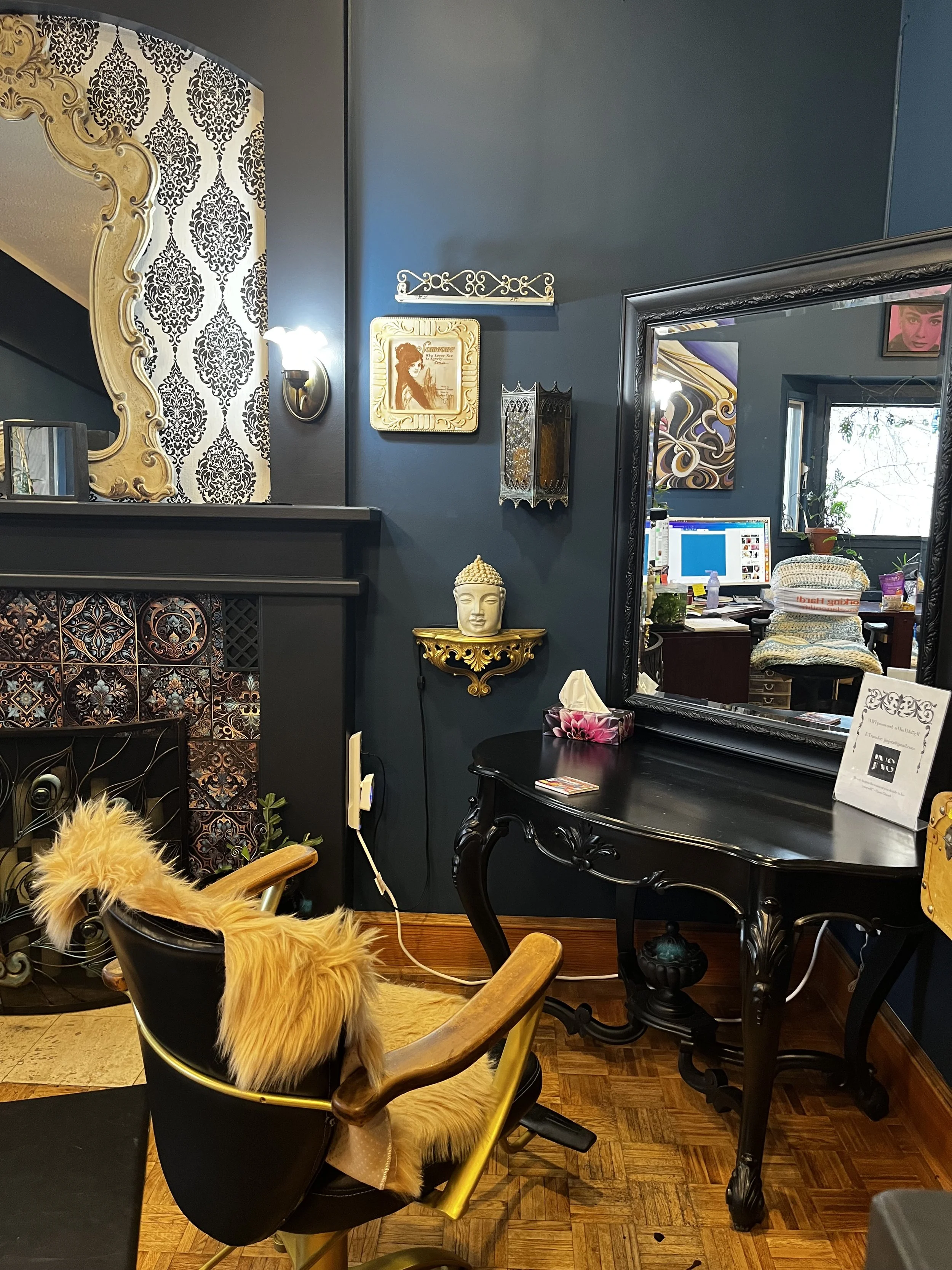 Luxurious black-and-gold salon interior with ornate damask wallpaper, decorative mirror, vintage artwork, Buddha statue, makeup desk, and salon chair.