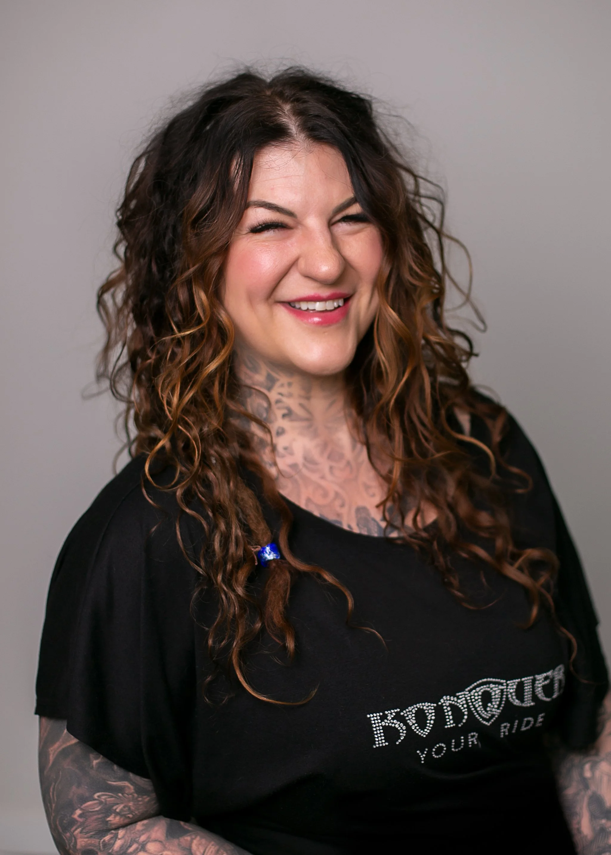 Smiling tattooed woman with long curly brown hair wearing a black T-shirt against a gray background