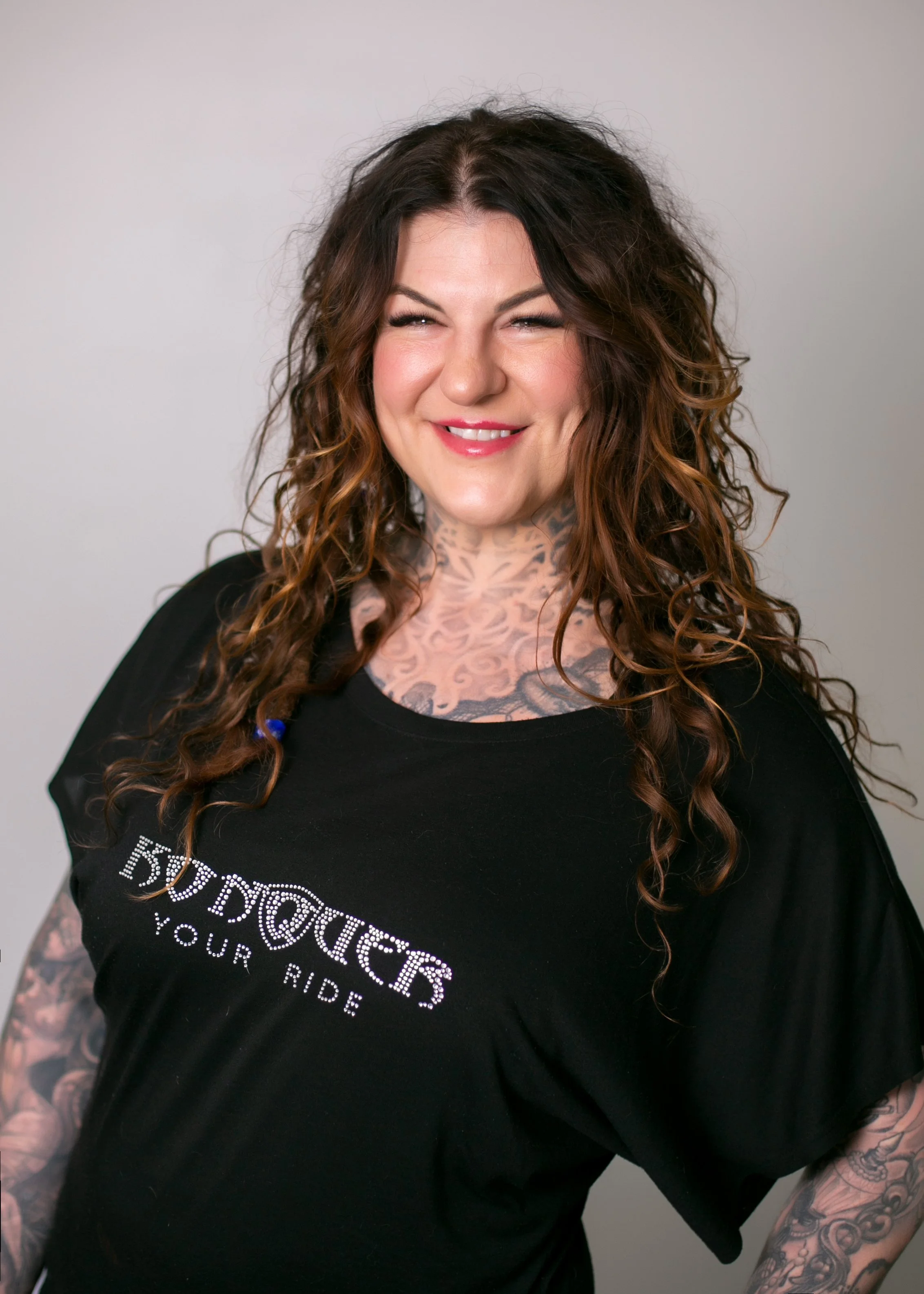 Smiling tattooed woman with long wavy brown hair wearing a black T-shirt against a plain background