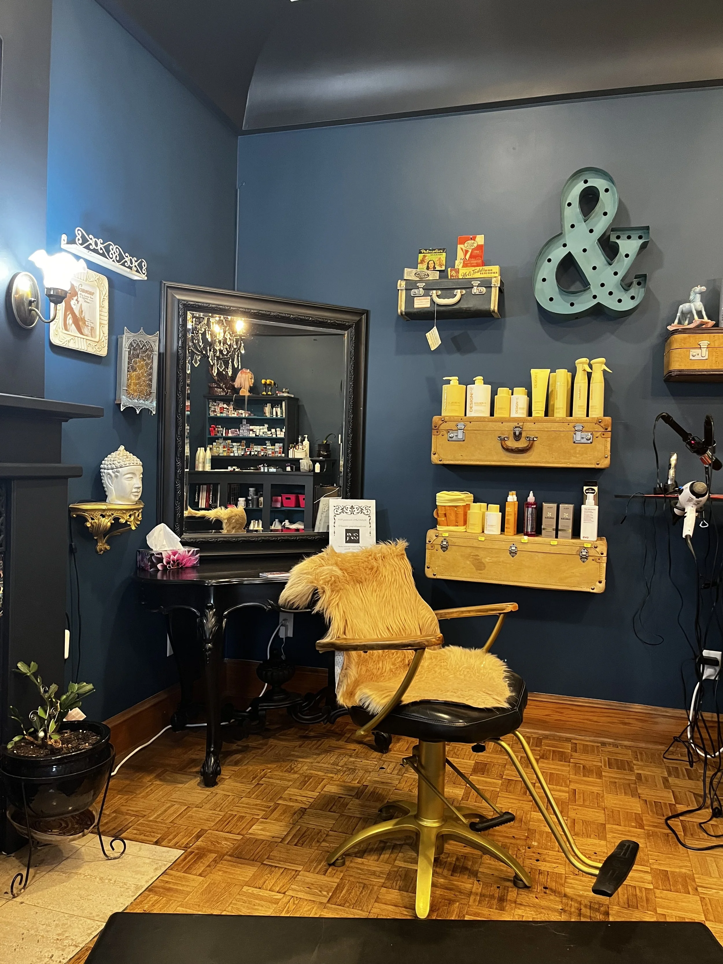 Vintage-inspired hair salon interior with a gold salon chair, dark blue walls, large mirror, hair products, decorative anchor, and hardwood floor