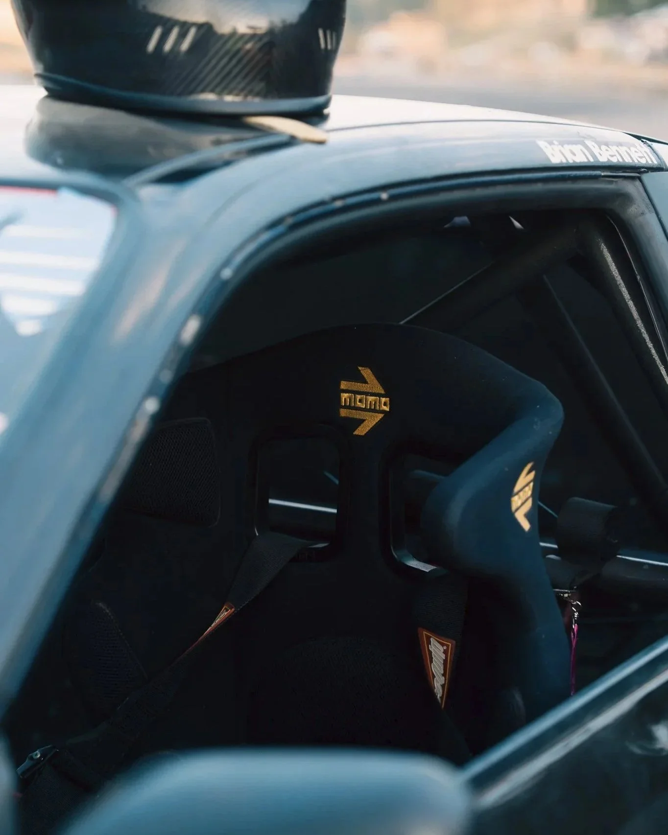 View from inside a car showing a racing helmet and a racing seat with Sparco branding. The car has a roll cage for safety, and a small black object, possibly a camera, is mounted on the dashboard.